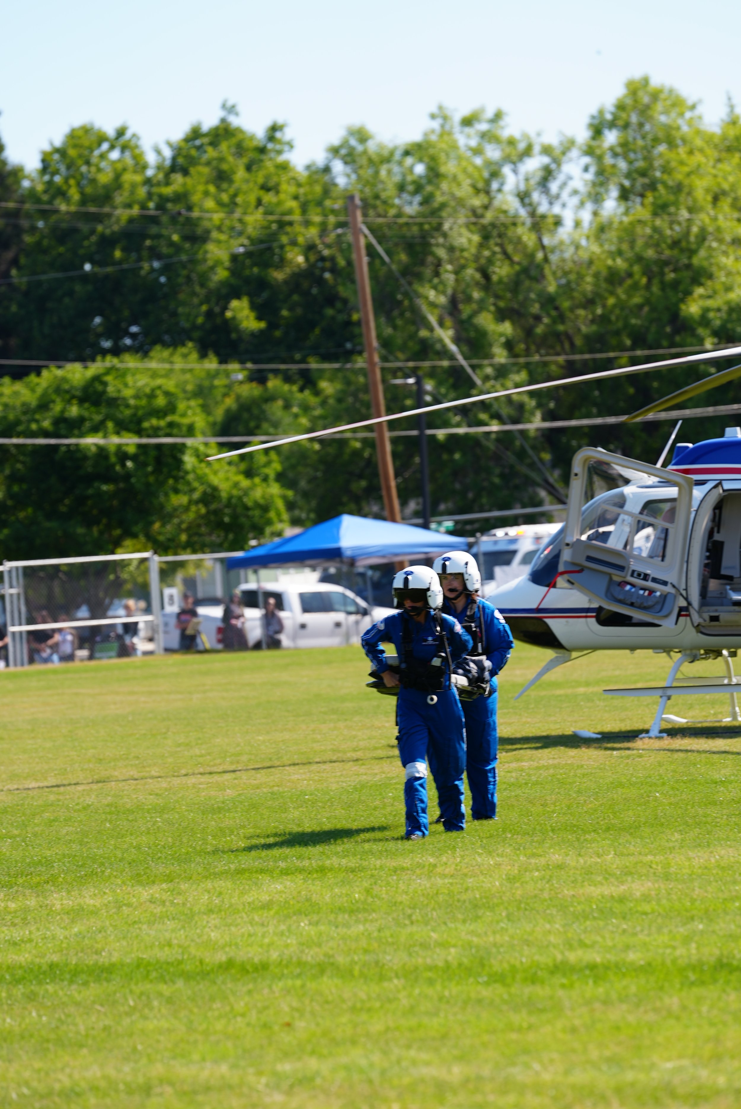 Two rescue personnel in blue uniforms and helmets walking on a grassy field near a helicopter, with trees, cars, and people in the background.