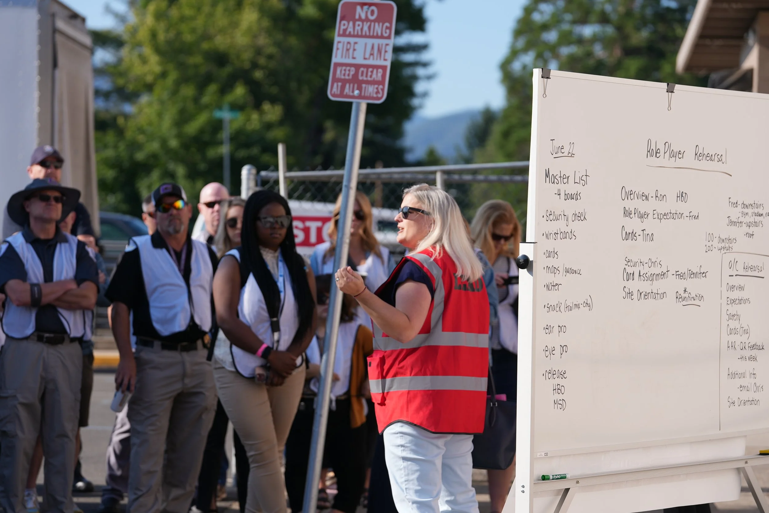A woman in a red vest giving a safety presentation to a group of people outside, with a whiteboard listing the agenda and a 'No Parking' sign in the background.