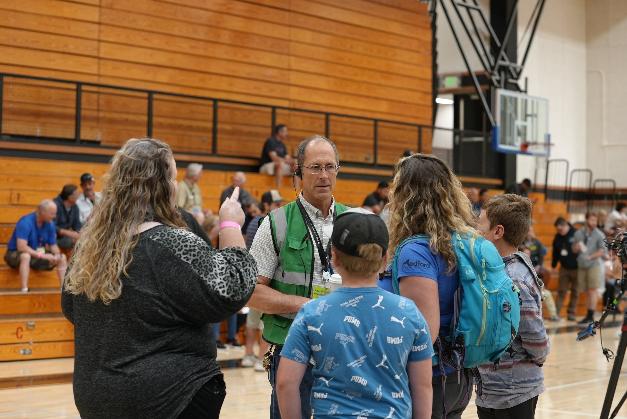 A group of people including a man with a green vest and headset, a woman in a blue shirt, and two children in casual clothing, are gathered in a gymnasium for a discussion or interview, with some seated in the bleachers in the background.
