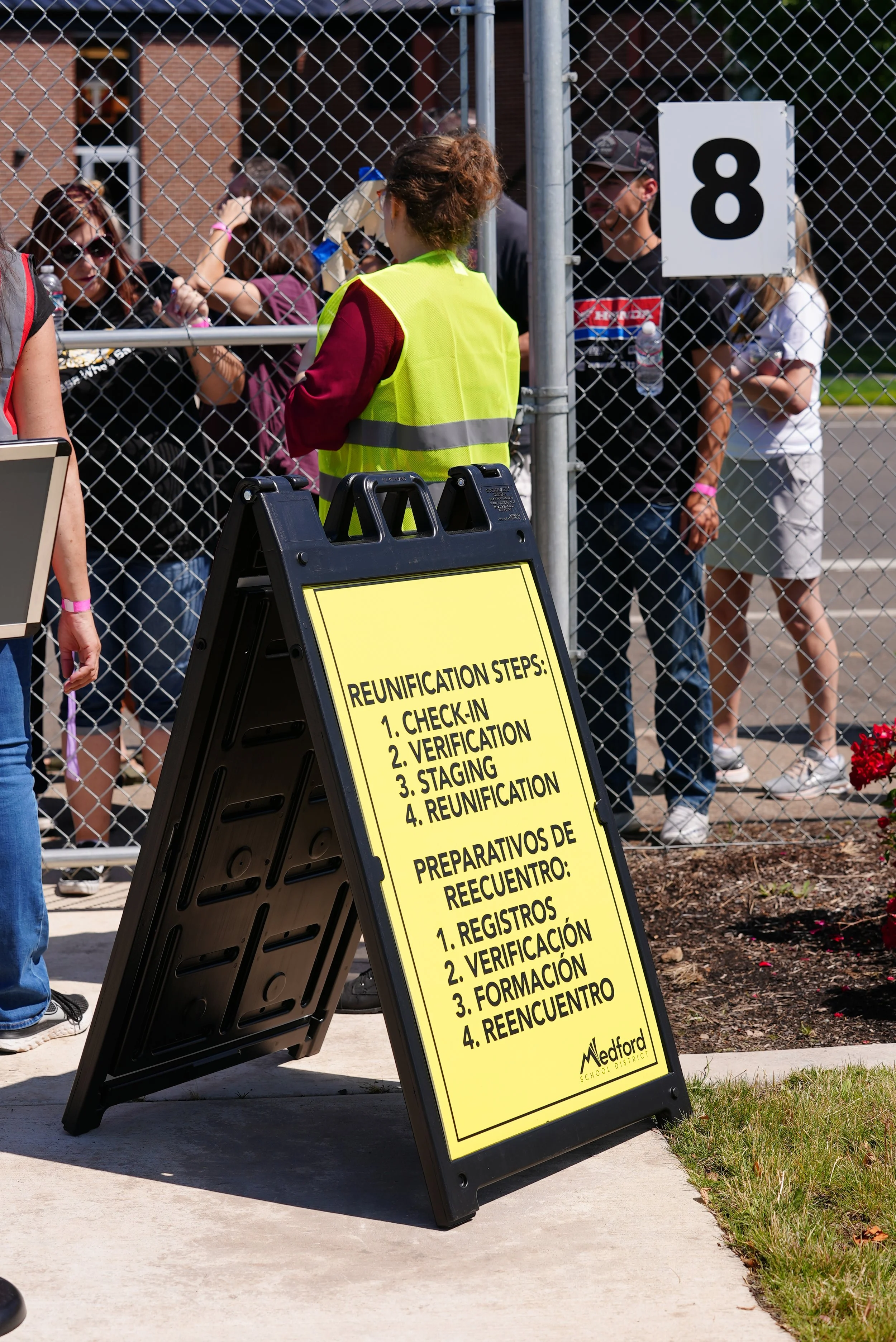 A group of people waiting behind a chain-link fence at a reunification event, with a sign listing steps for reunification and verification.