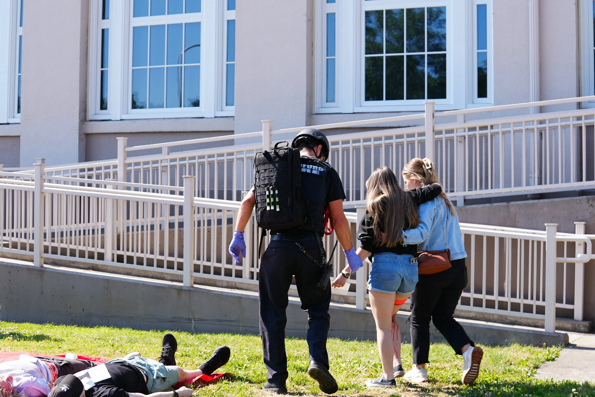 A police officer assisting a young girl with a leg injury while two other young girls support her. A person is lying on the grass nearby.