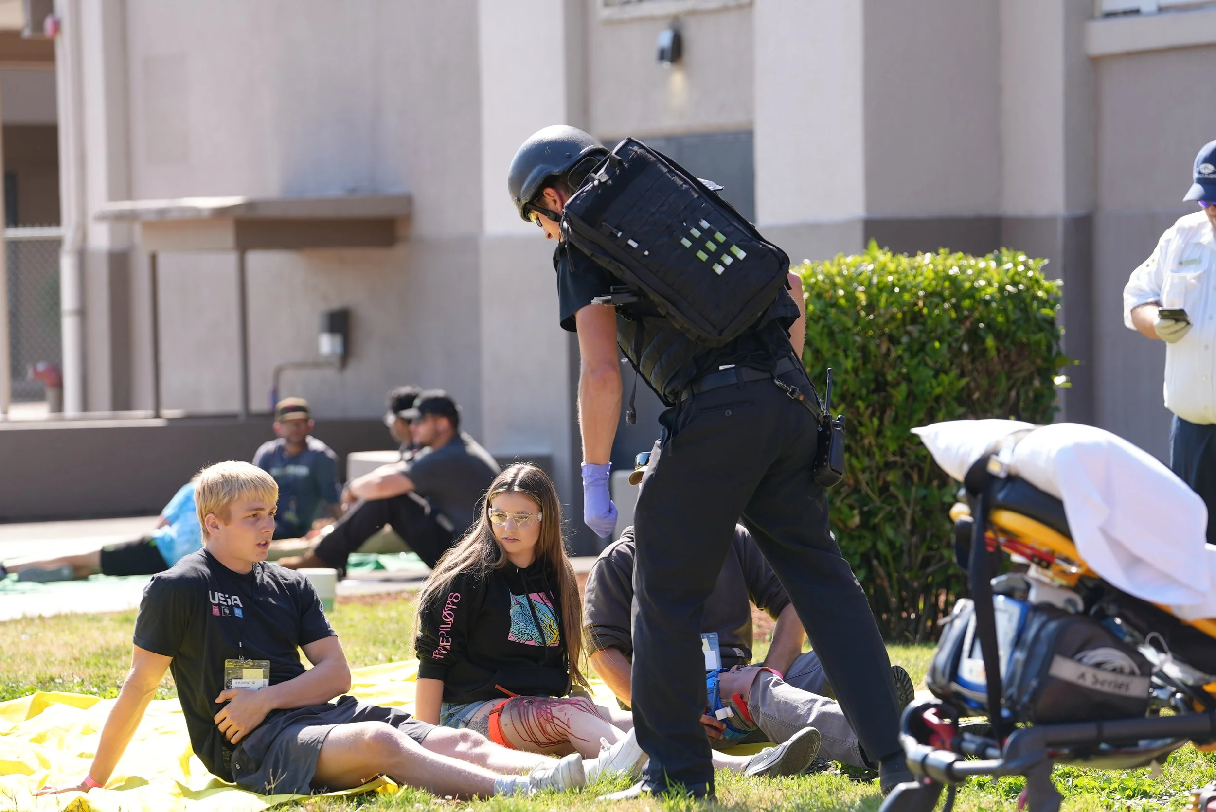 Emergency responders providing first aid to a group of young people sitting on a yellow blanket outside a building.