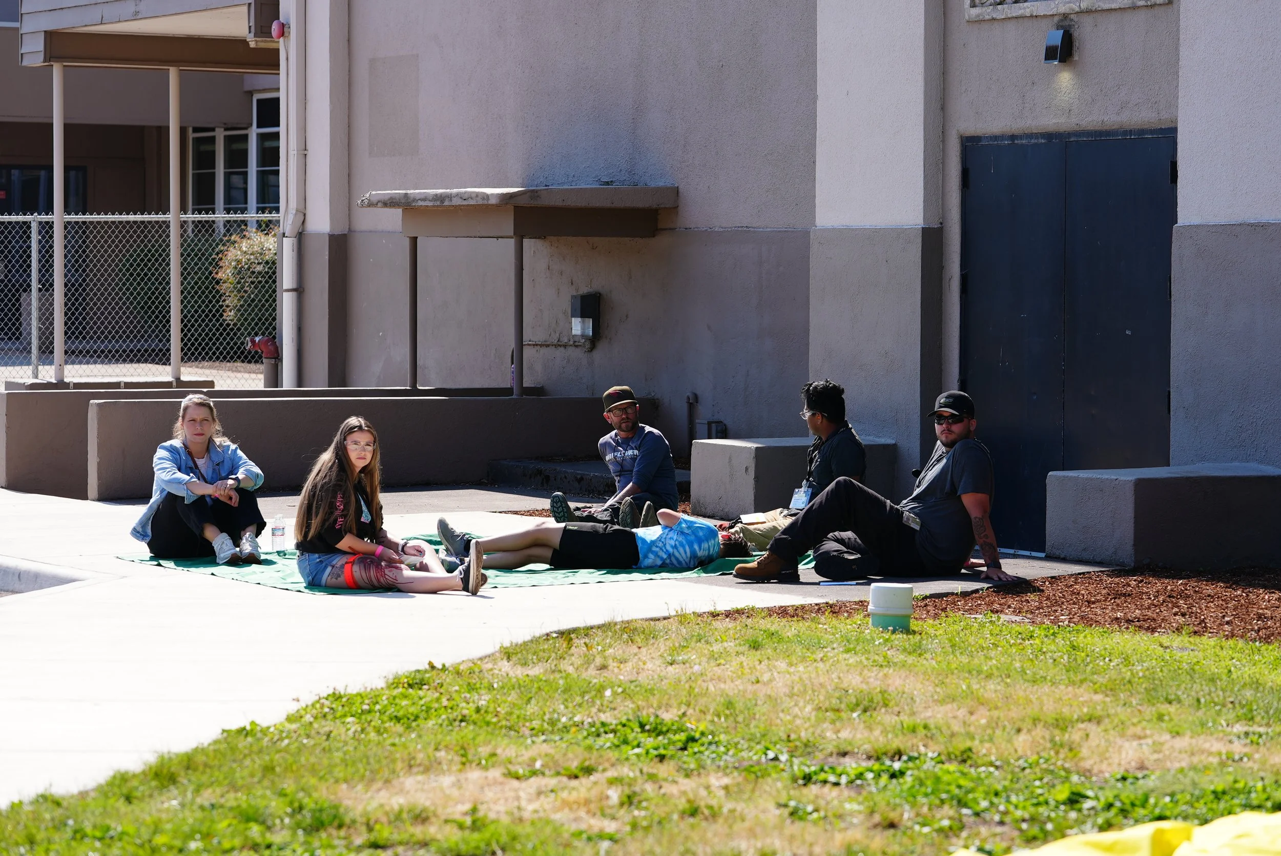 Group of six young people sitting and lying on a blanket outside a beige building with a door and some concrete structures, on a sunny day.