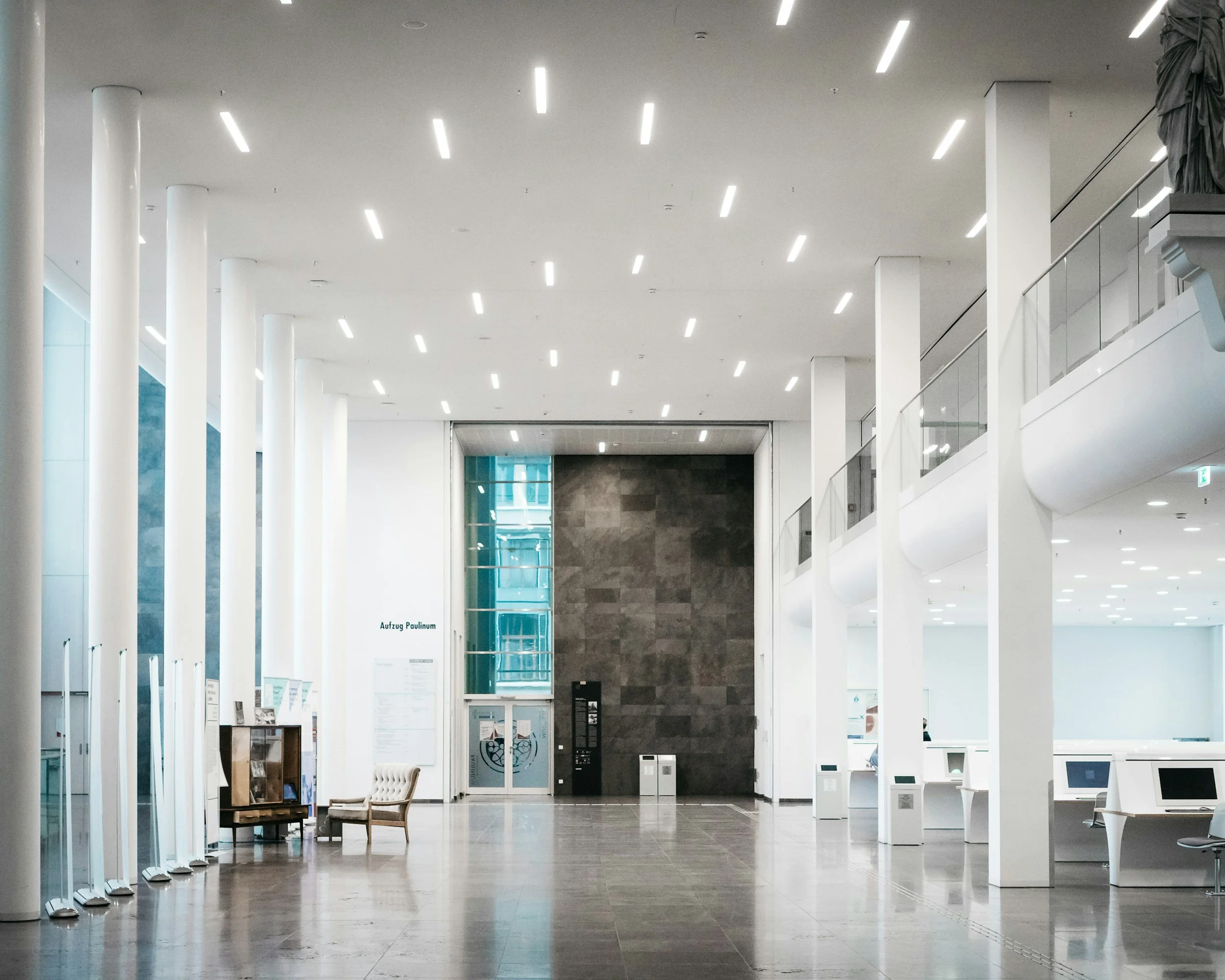 Empty modern lobby with white walls, tall white pillars, and ceiling lights, featuring some chairs, a bookcase, a vending machine, and digital kiosks.