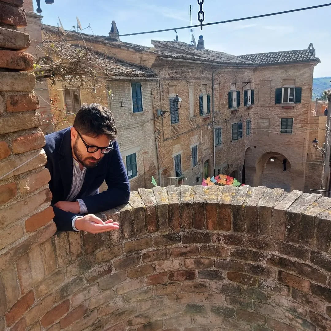Uomo con occhiali, barba e giacca appoggiato a un muro di mattoni, guarda cercando qualcosa sopra.