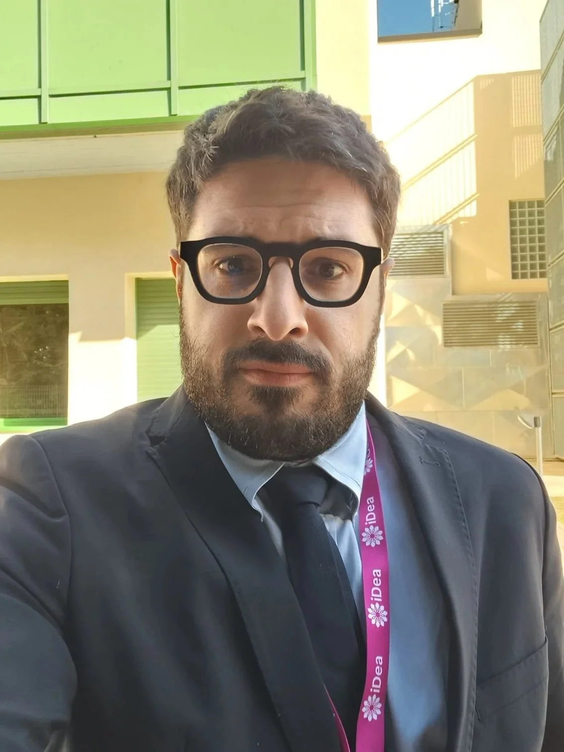 Uomo con barba e occhiali grandi, vestito in abito nero e cravatta, posa per un selfie con edificio moderno sullo sfondo.