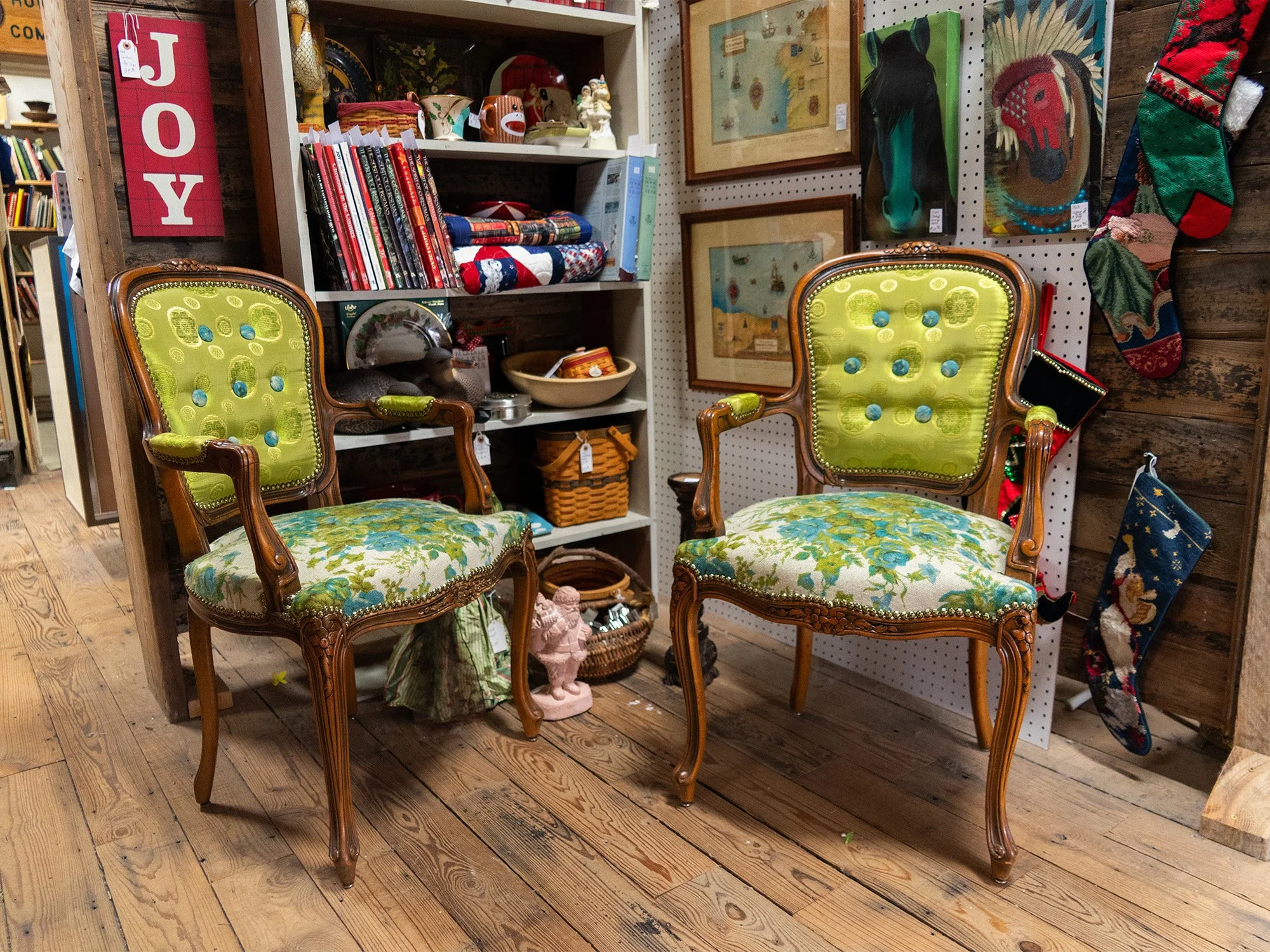 Two vintage wooden chairs with green upholstery and floral fabric on the seats are placed in front of shelves and a pegboard wall decorated with vintage artwork and Christmas stockings.