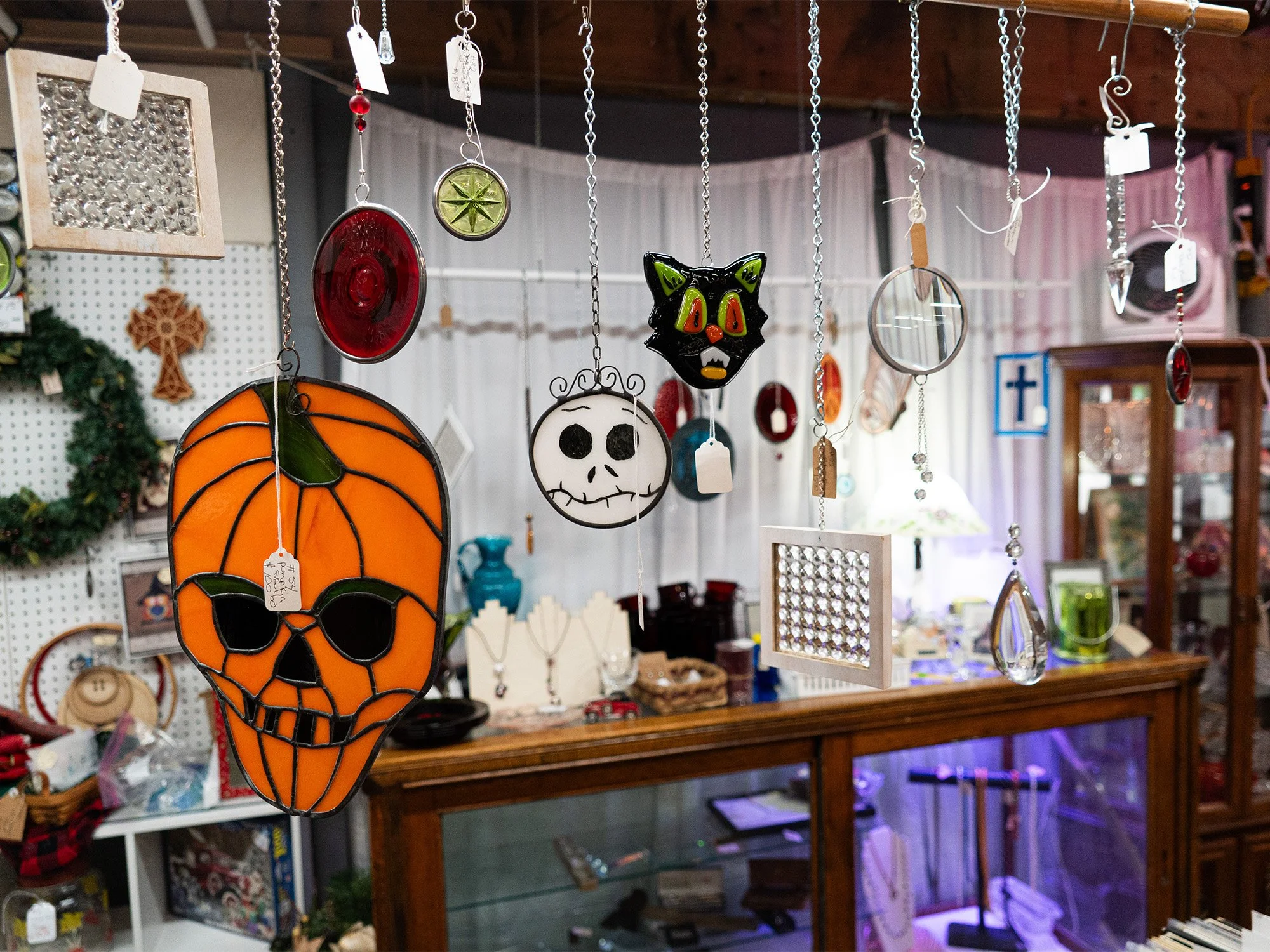 Hanging Halloween-themed glass decorations, including a pumpkin skull, a white skull, a black cat face, and various circular pieces, displayed in a gift shop with wooden furniture and festive decorations.