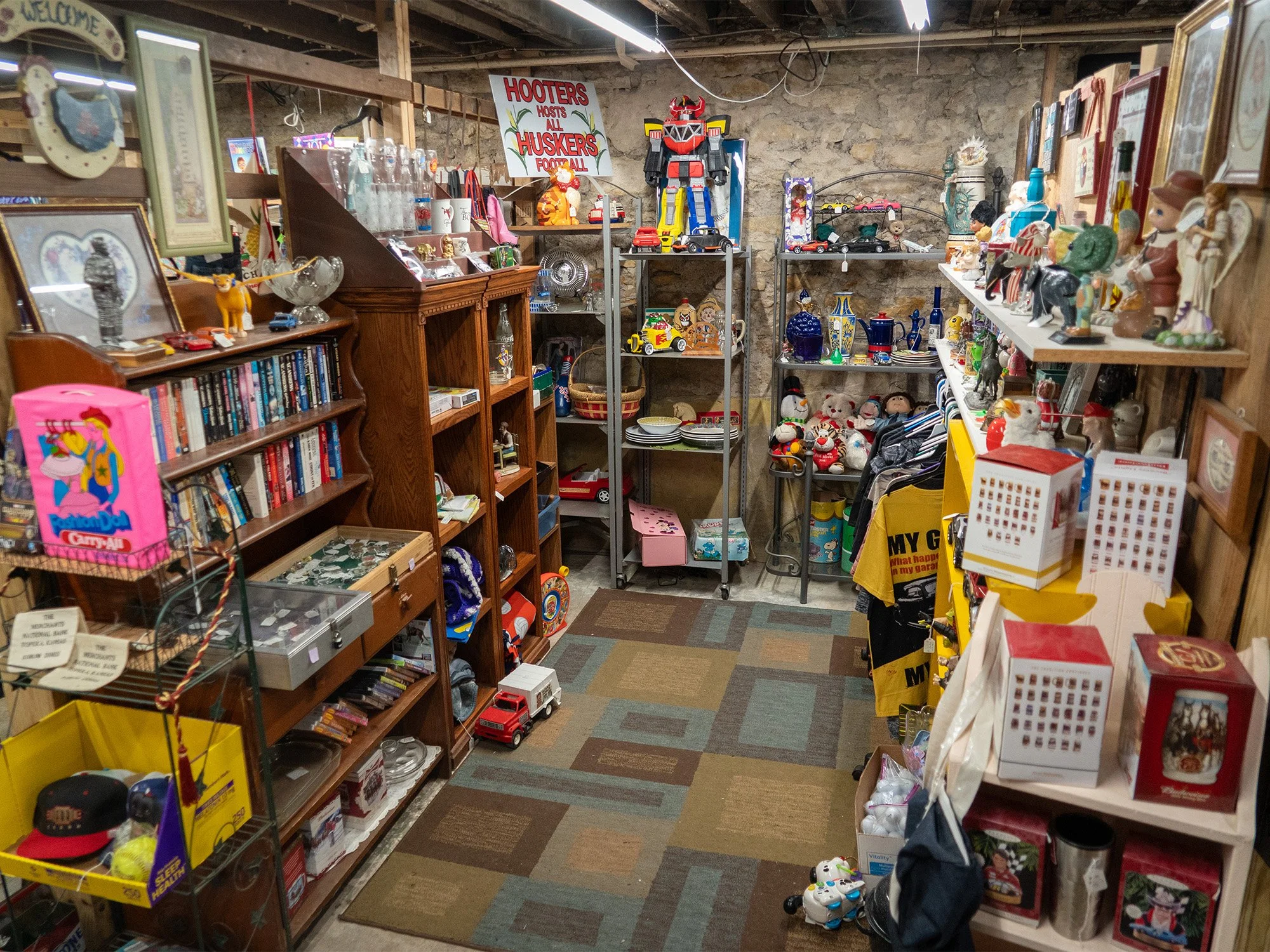 A thrift shop filled with various collectible items, toys, and decorative objects on shelves, with a stone wall background and a colorful carpet on the floor.
