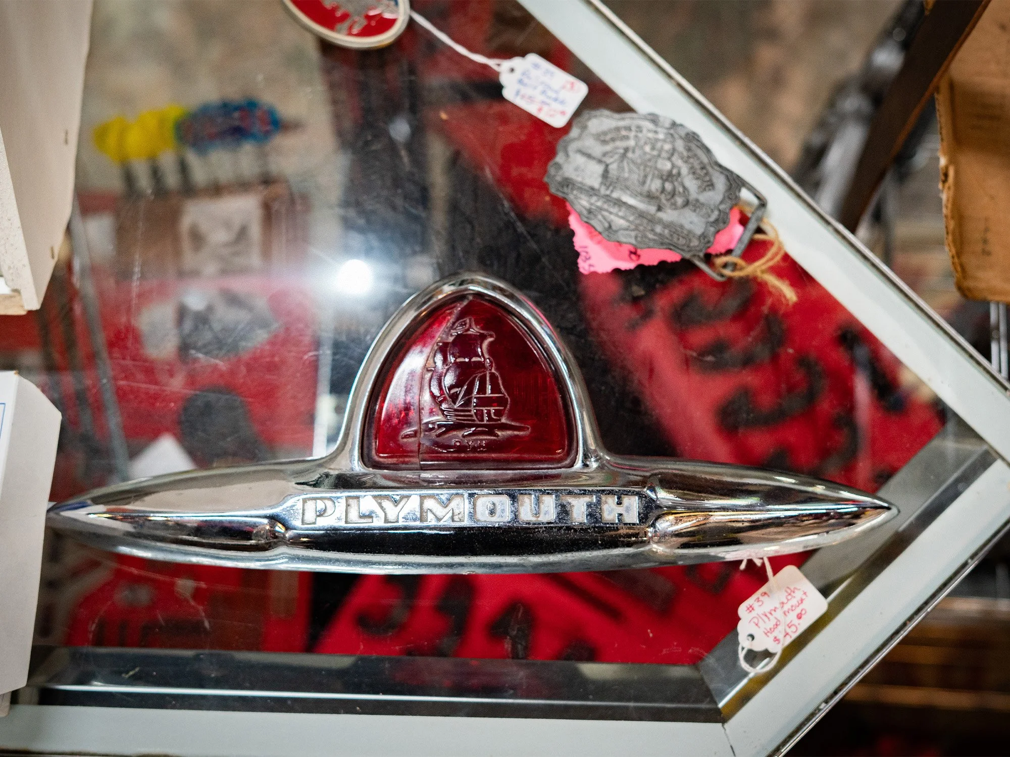 A vintage Plymouth car emblem on a glass display case with a red background. The emblem features a ship at the top and the word 'PLYMOUTH' engraved below. There are price tags on the case indicating it is an item for sale.