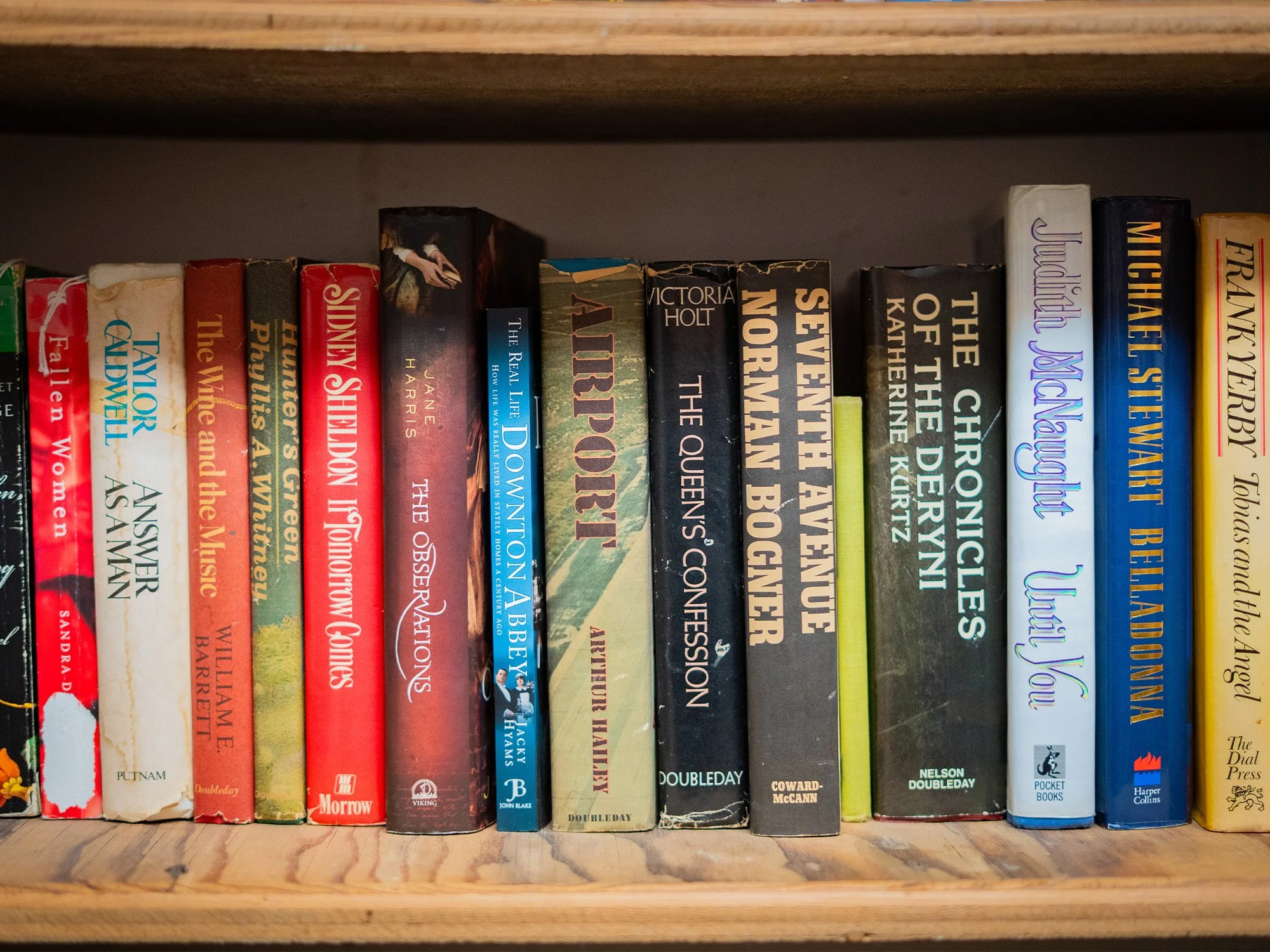 Row of hardcover and paperback books on a wooden bookshelf, with titles including 'The Queen's Confinement,' 'The Night Before,' 'The Chronicle of the Deryn,' and 'Very You'.