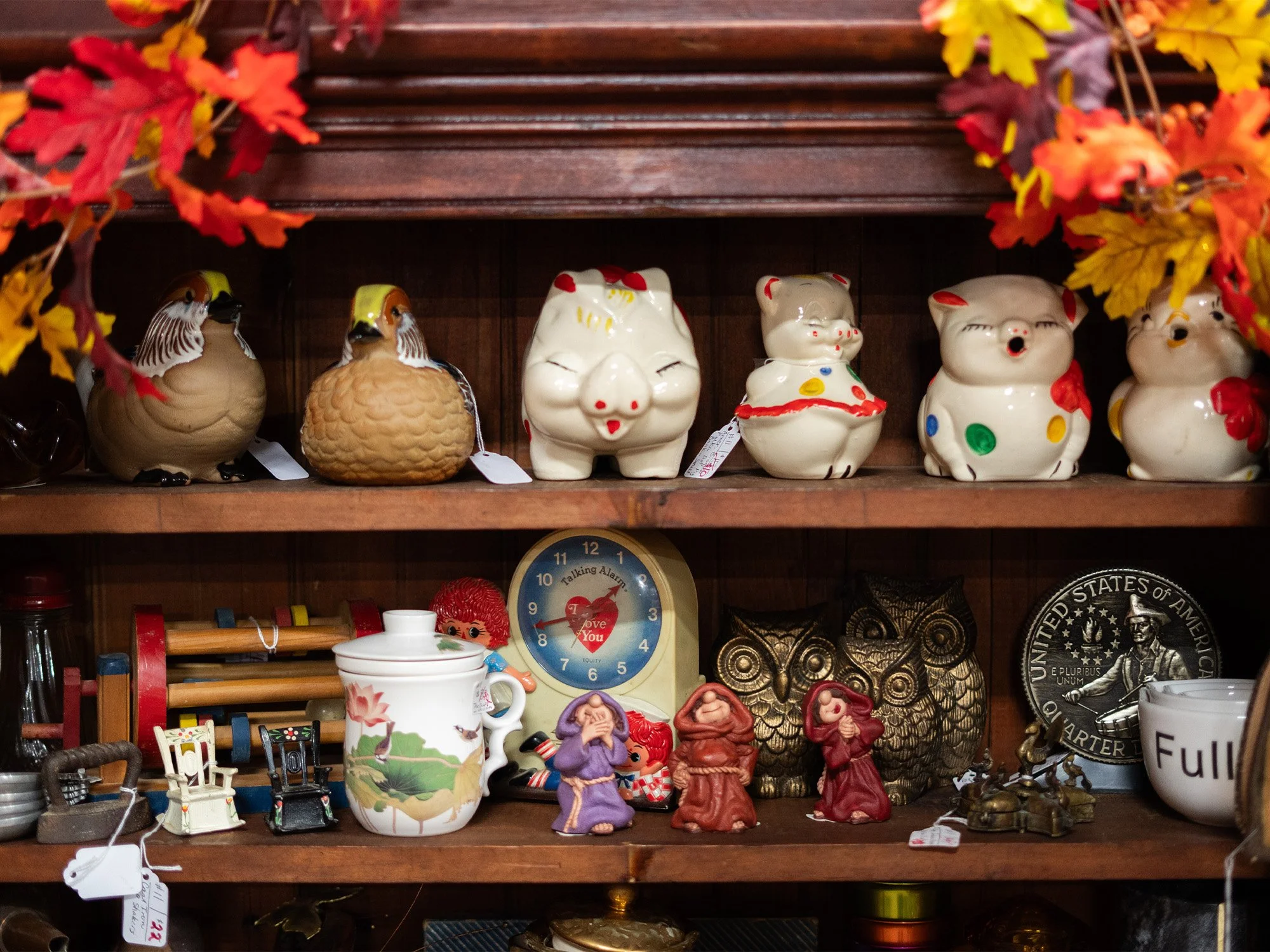 Shelves filled with various decorative items, including ceramic piggy bank figurines, Owl figurines, small figurines of monks, a clock, a teapot, and other miscellaneous collectibles. The top shelf has piggy bank figurines and the lower shelf has owl