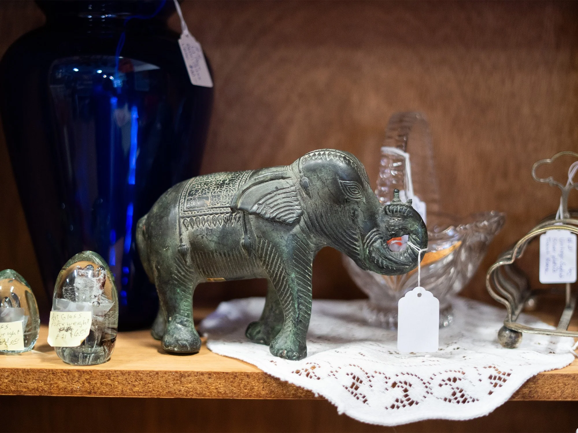 A metal elephant figurine with intricate carvings, placed on a lace cloth, surrounded by glass decorative objects on a wooden shelf.