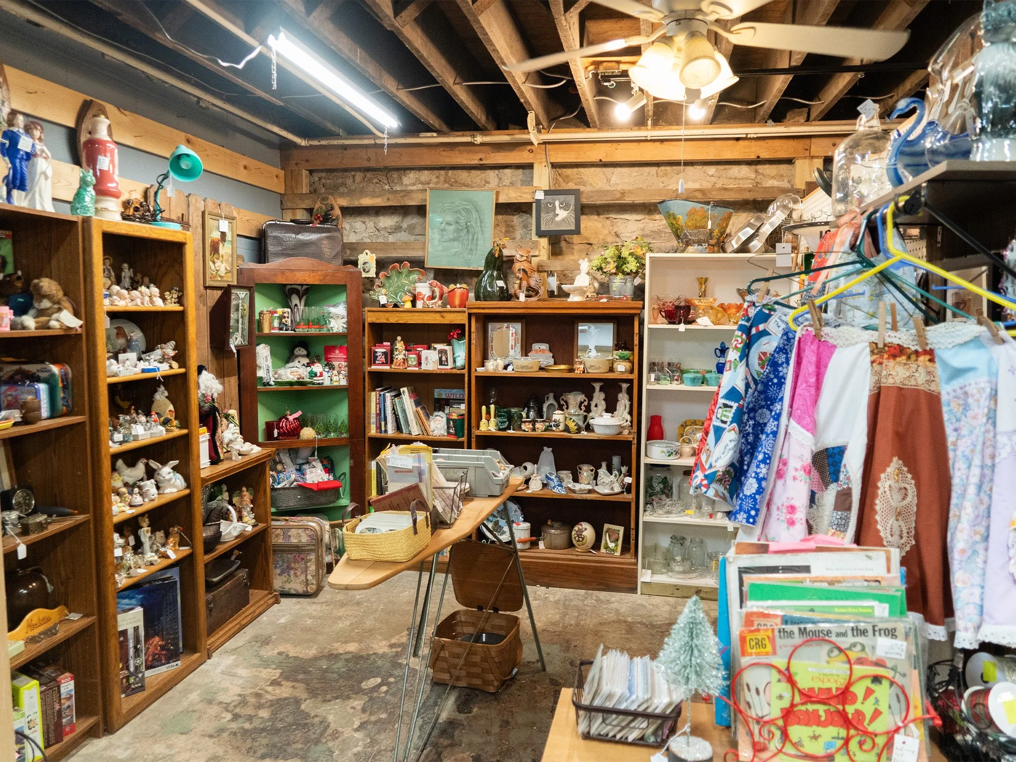 Interior view of a thrift store or antique shop with shelves filled with various collectibles, figurines, and dishes, clothing on a rack, and a wooden table with small items; exposed wooden beams on ceiling and framed artwork on the back wall.