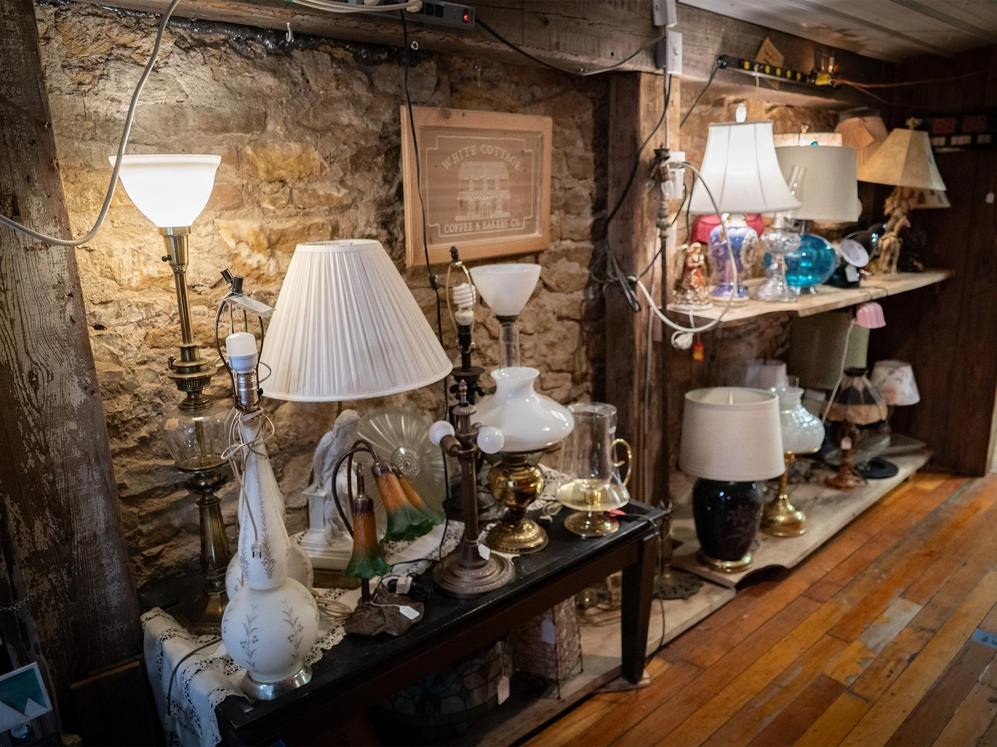 Various lamps and decorative glassware displayed on shelves and tables against a stone wall in a rustic setting.