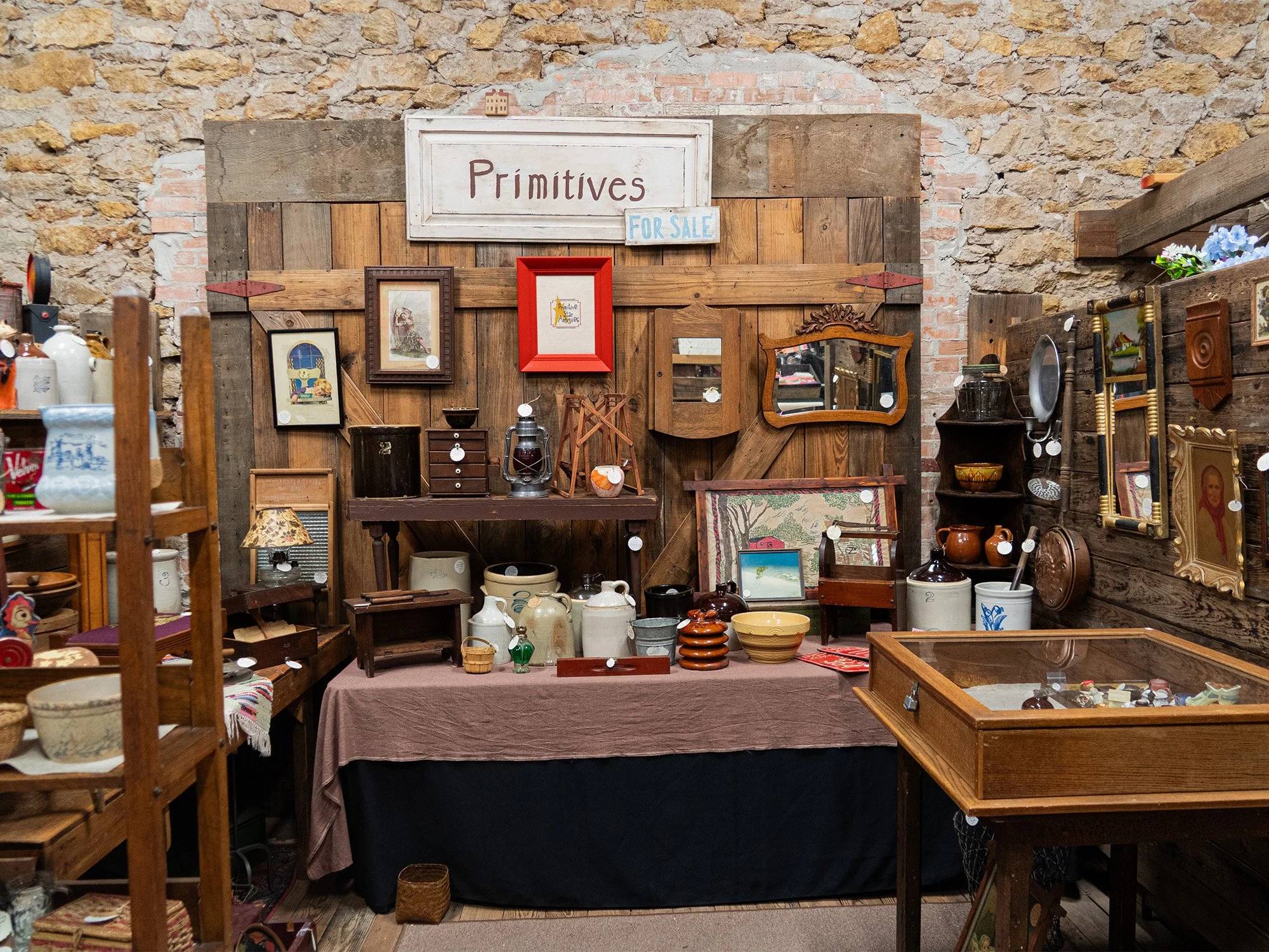 A display of vintage and antique items at a thrift or antique shop, with a rustic brick and wood wall background, labeled 'Primitives' and 'For Sale' signs, various picture frames, ceramics, glassware, and small furniture pieces arranged on tables an