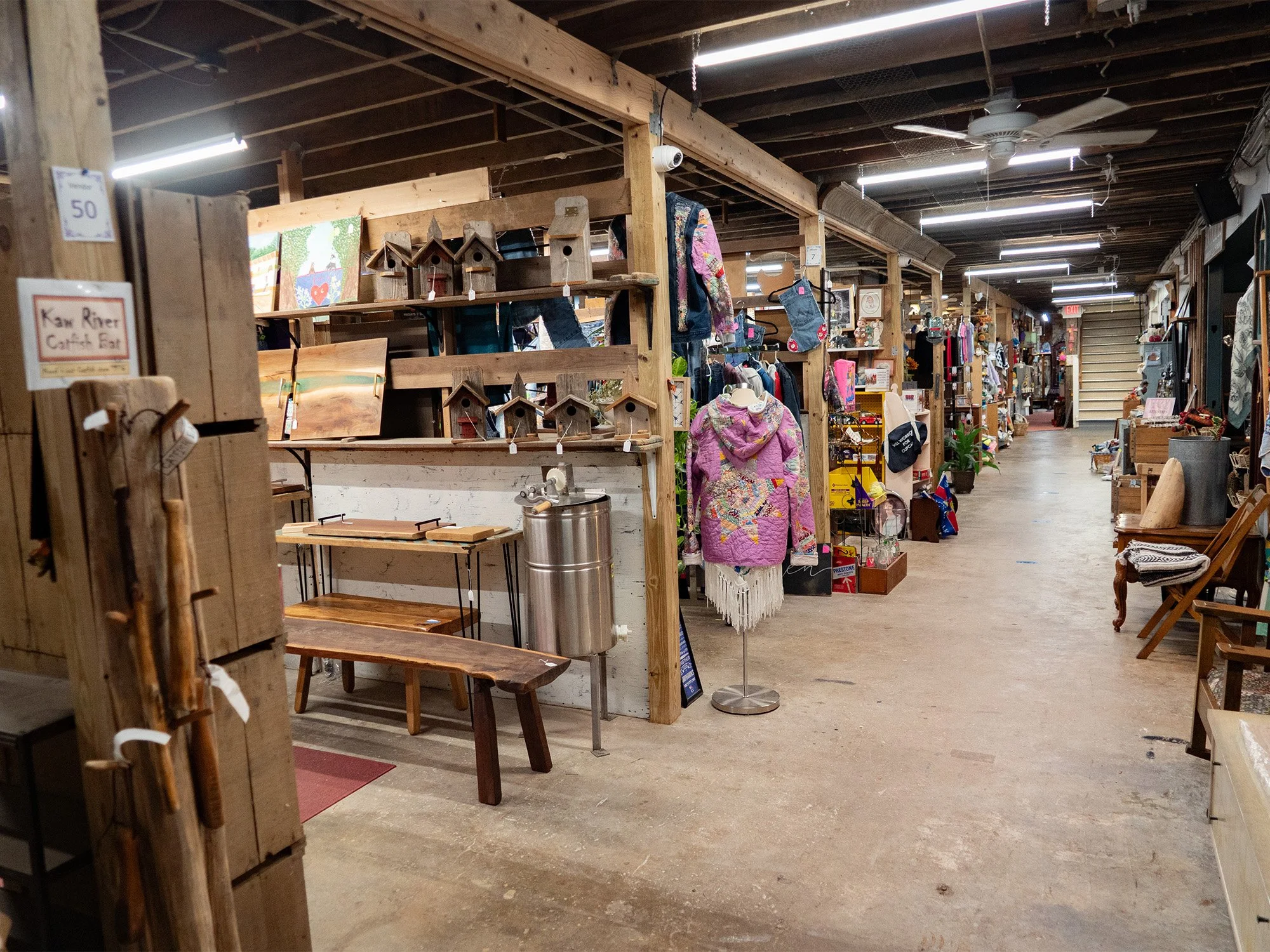 Interior of a thrift store with wooden shelves displaying paintings, birdhouses, and clothing on racks. A pink quilted jacket is on a mannequin. The store has exposed wooden beams, ceiling fans, and a concrete floor, with several aisles filled with v