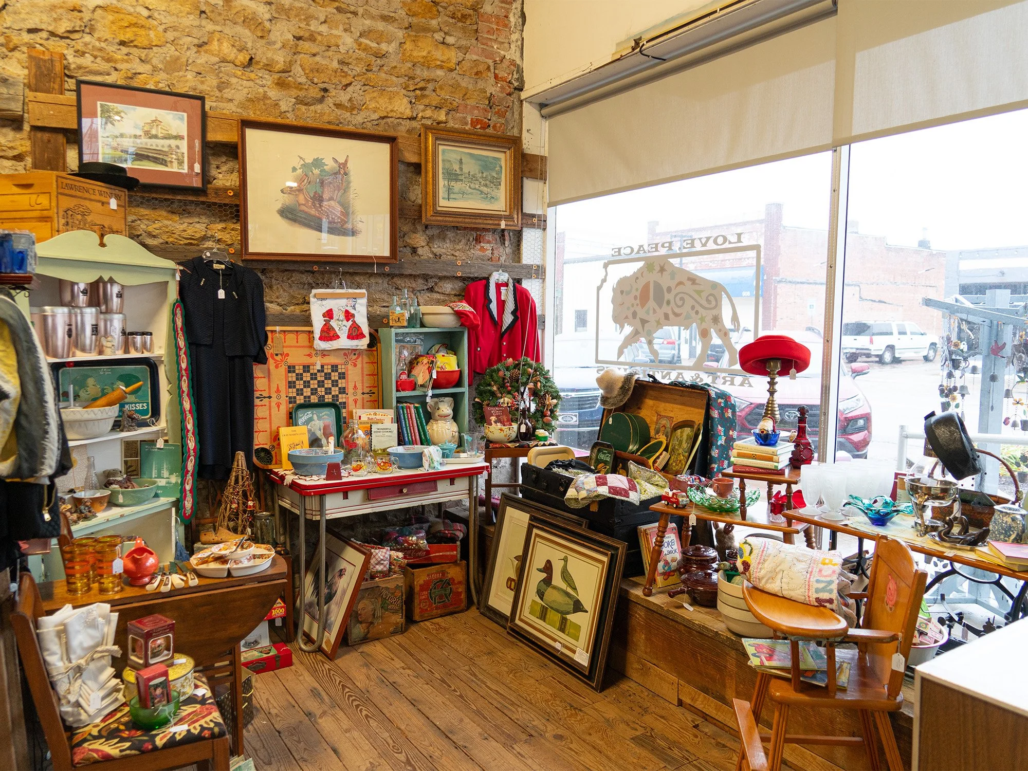 Interior of a vintage thrift store with various antiques, artwork, and clothing displayed on shelves, tables, and walls, with a large window showing parked cars outside.