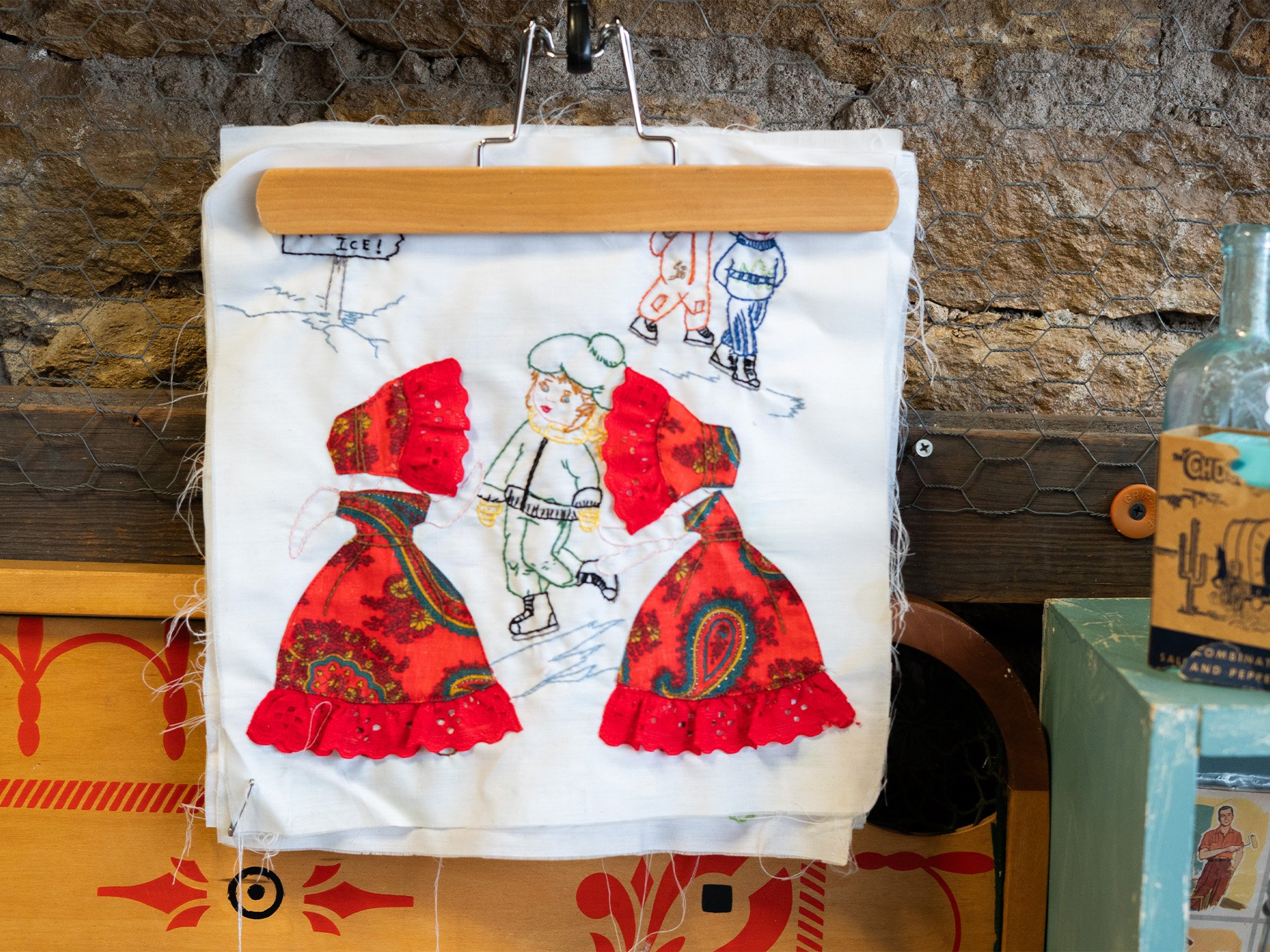 Embroidery hoop with a fabric piece featuring embroidered and sewn Christmas-themed figures, including a boy ice skating, two women in colorful coats with red ruffles, and a child with a green hat.