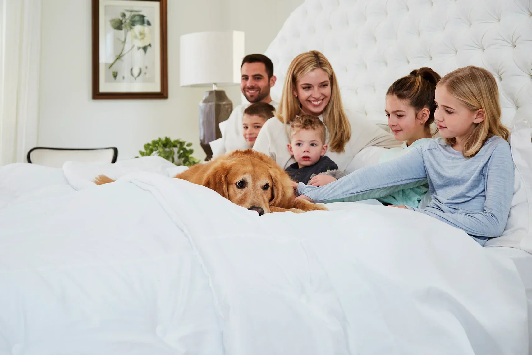 Family with children and a dog on a bed in a well-lit bedroom.