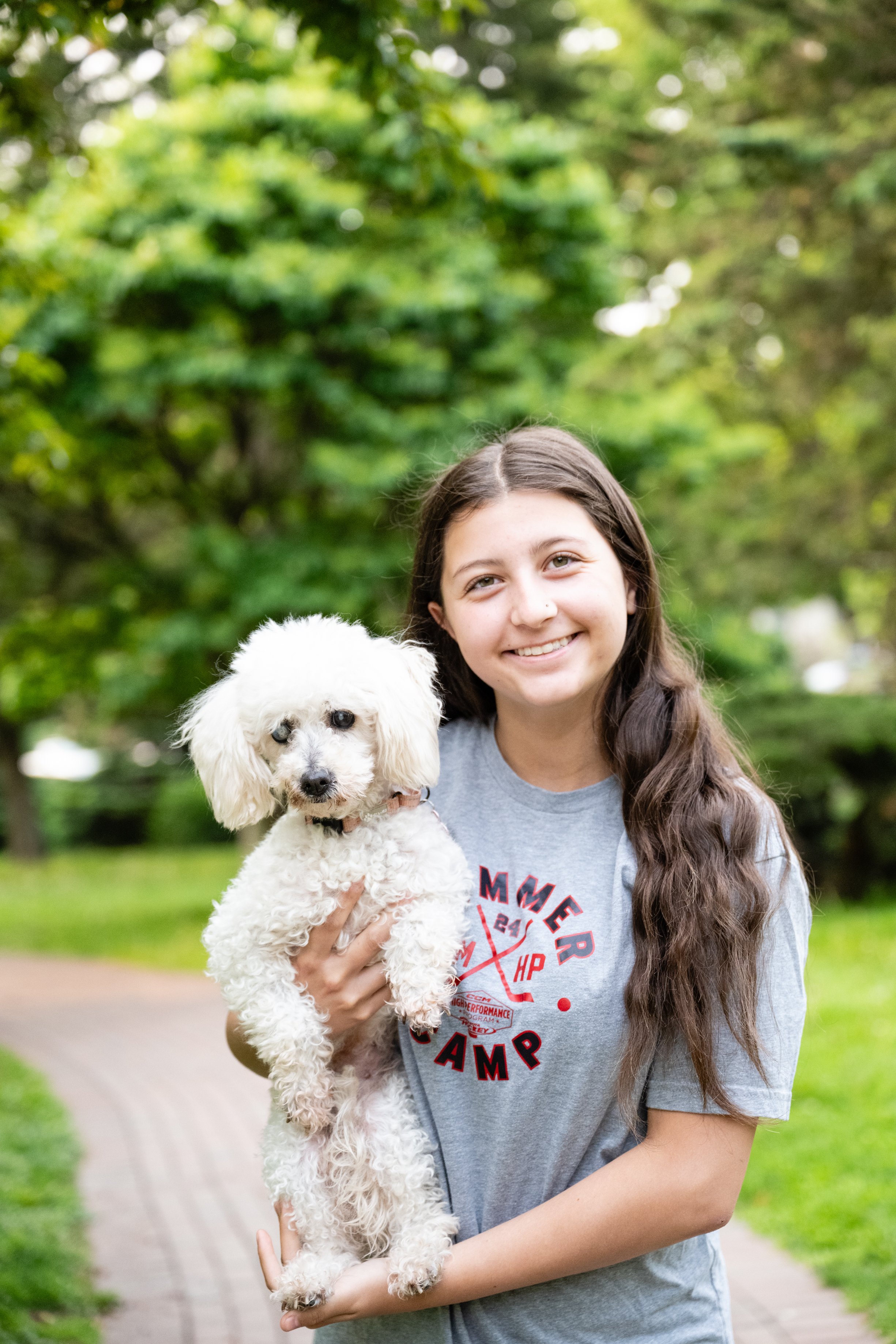 A girl with long brown hair smiling and holding a small white poodle outdoors with green trees and a brick pathway in the background.