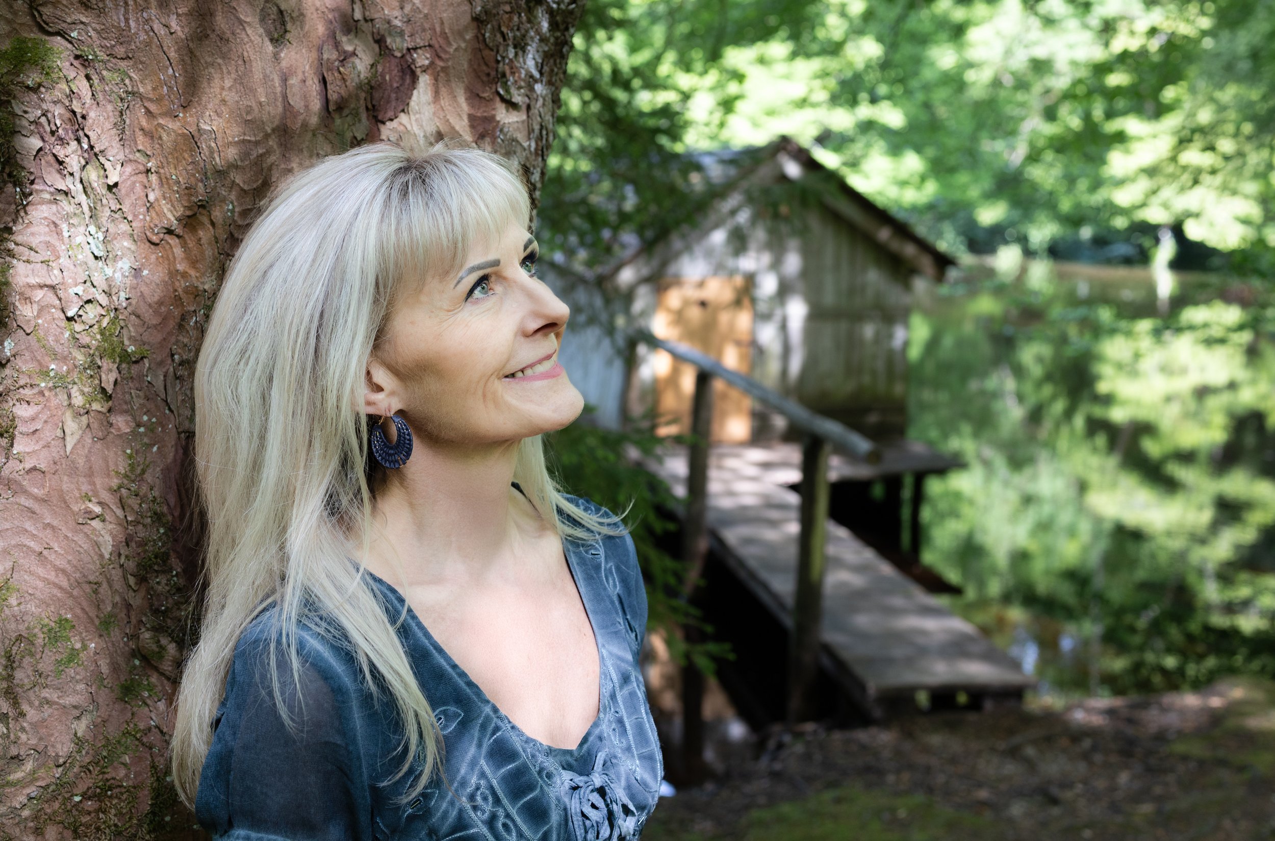 Eine lachende Frau mit blonden Haaren, die an einem Baum im Grünen lehnt, im Hintergrund eine kleine Holzbrücke und ein Holzhaus in einem Wald.