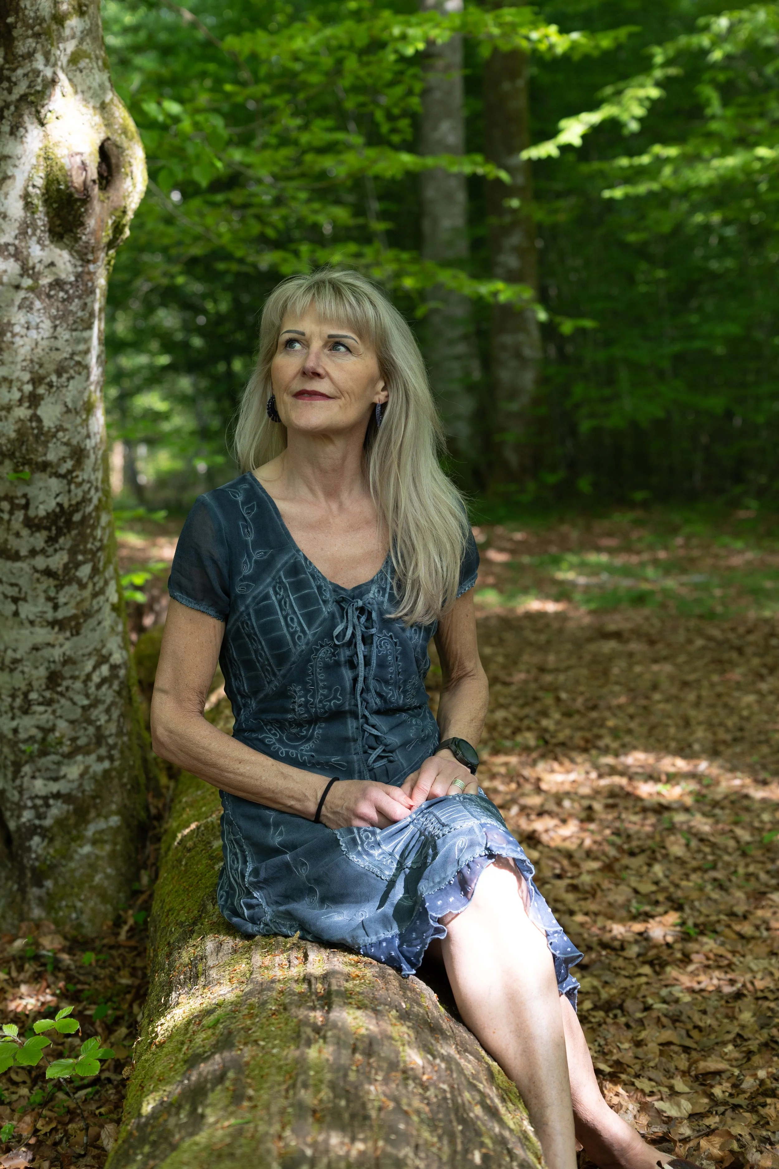 Eine ältere Frau sitzt auf einem Baumstamm in einem grünen Wald, trägt ein dunkelblaues Kleid und schaut nach oben.