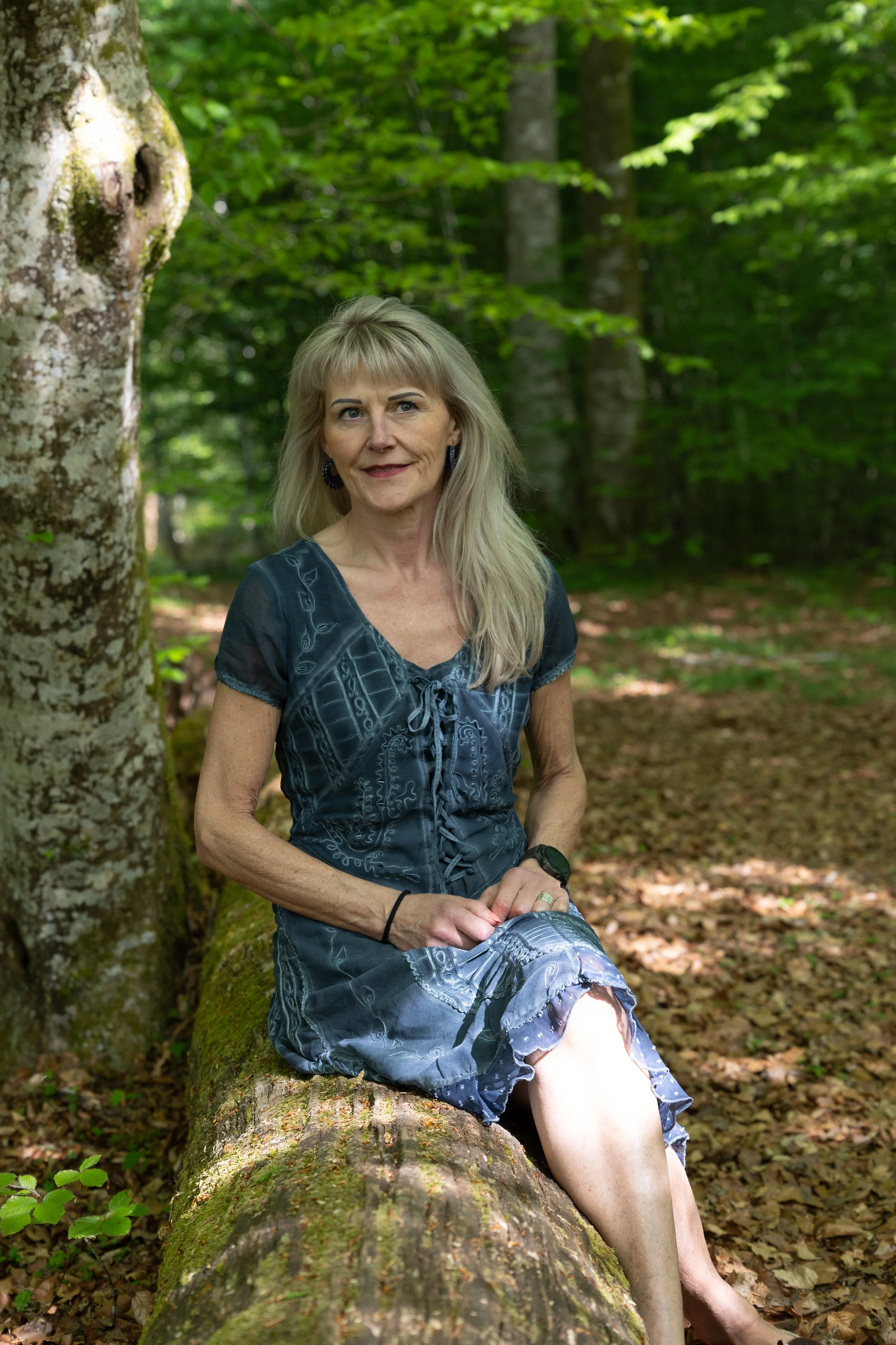 Eine ältere Frau mit blonden Haaren sitzt auf einem Baumstamm im Wald, umgeben von grünen Bäumen und Laub.