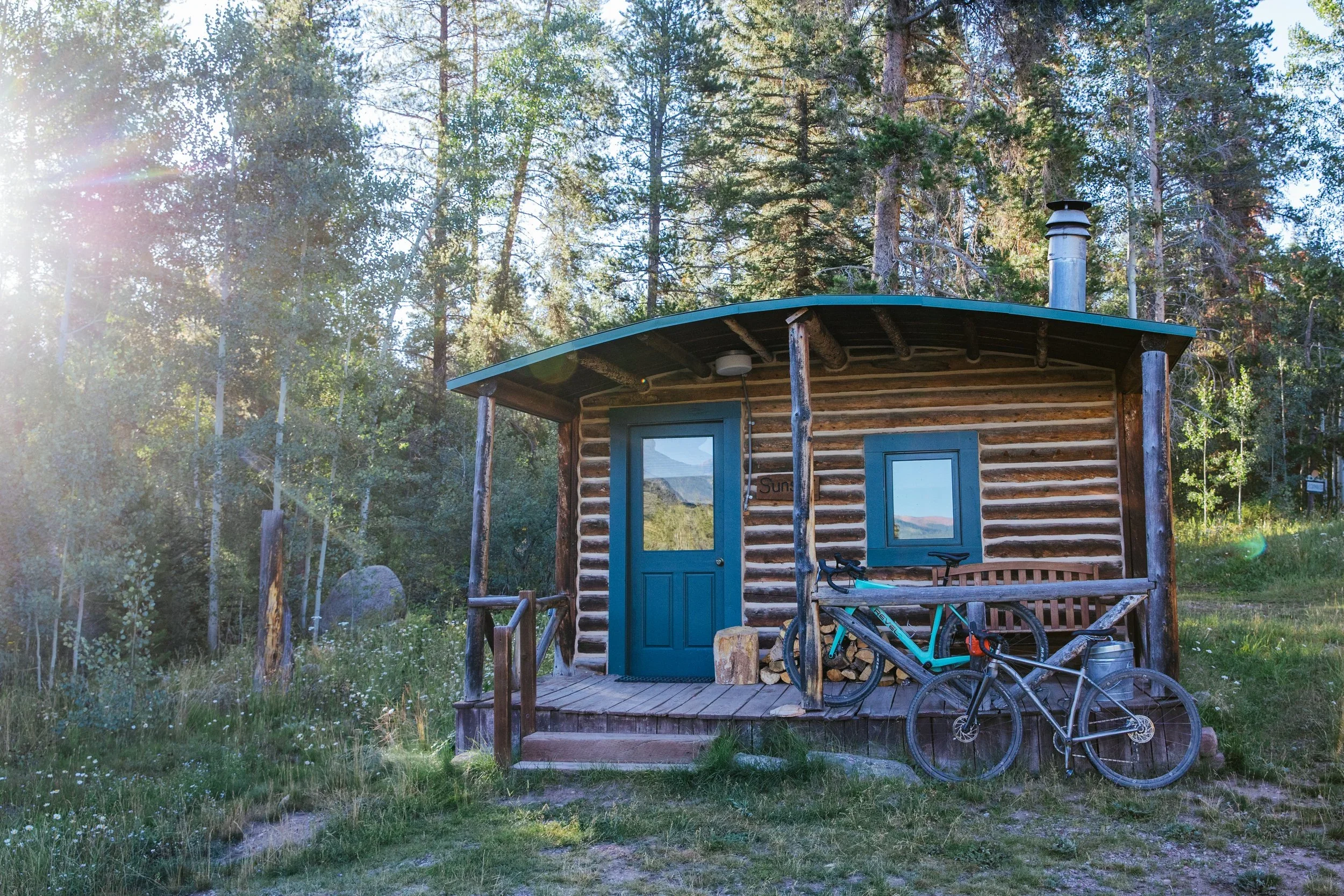 Small log cabin with a blue door and window, surrounded by trees, with a mountain view reflected in the window. There are two bicycles parked on the porch.