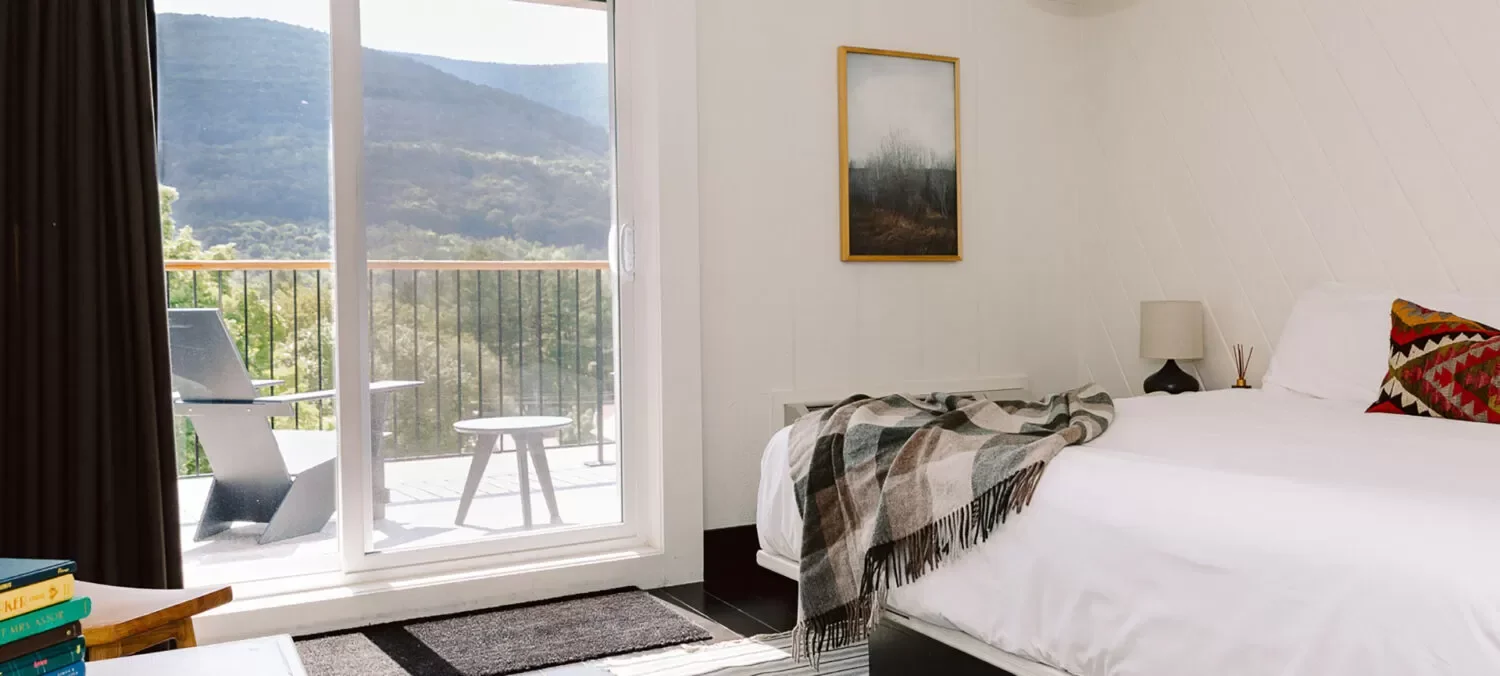 A bedroom with a bed, pillow, blanket, small lamp, artwork on the wall, sliding glass door with a balcony, and mountain view outside.