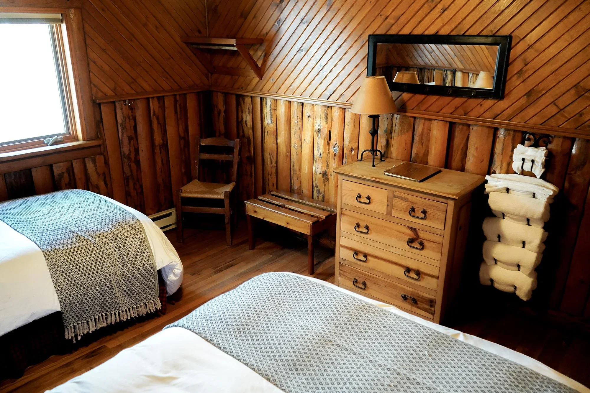 Room with wooden paneling, two beds with patterned bed coverings, a window, a wooden dresser with a lamp and a mirror above, a stack of rolled towels, a chair, and a wooden bench.
