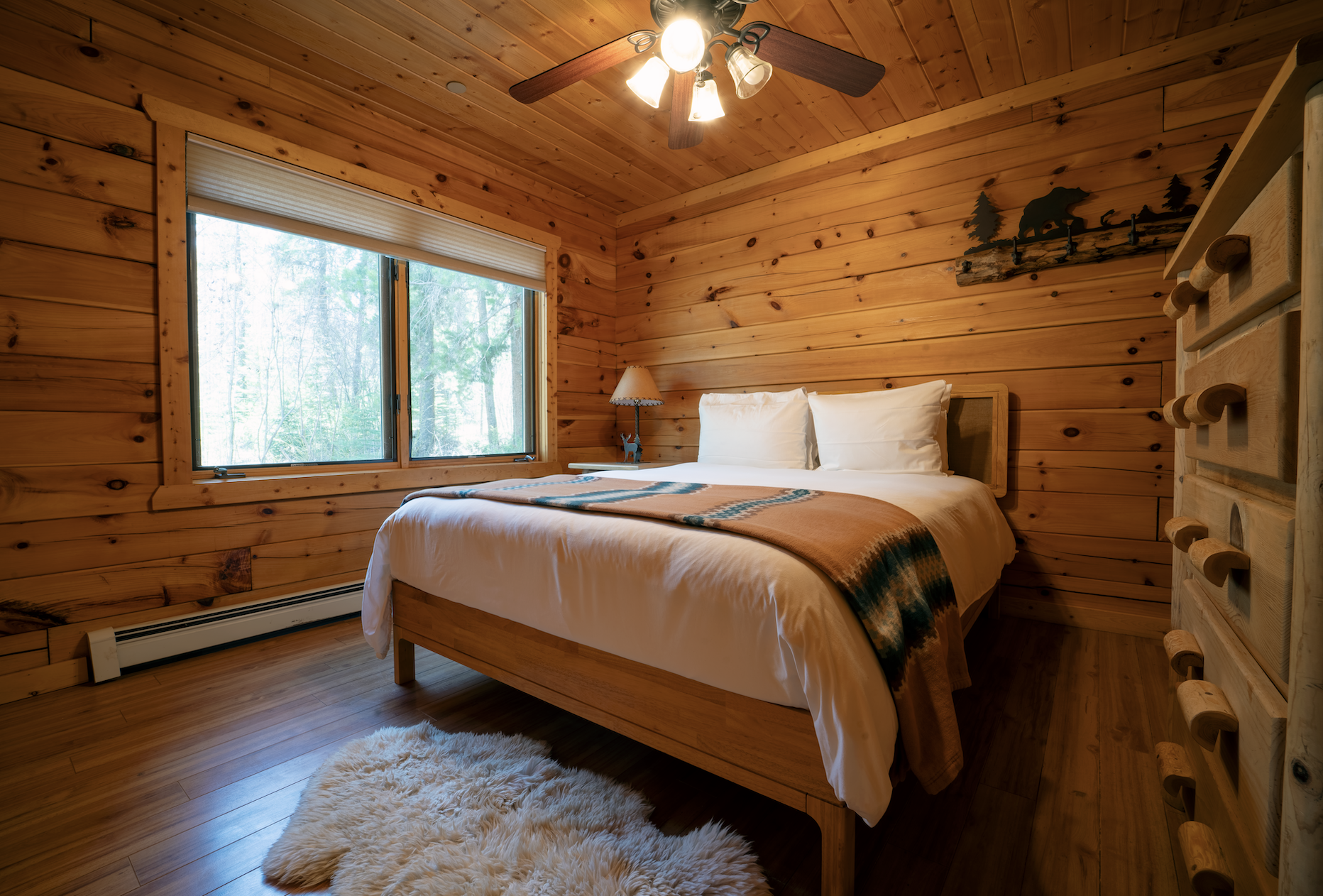 A cozy bedroom with wood-paneled walls, a window looking out to trees, a bed with white bedding and an orange-brown blanket, a small nightstand with a lamp, and a fluffy white rug on the wooden floor.