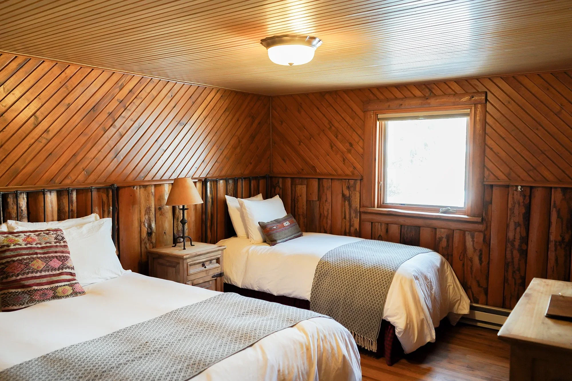 A rustic bedroom with two beds featuring white linens and patterned throws, wooden paneling on the walls, a small bedside table with a lamp between the beds, a window letting in natural light, and a wooden dresser.