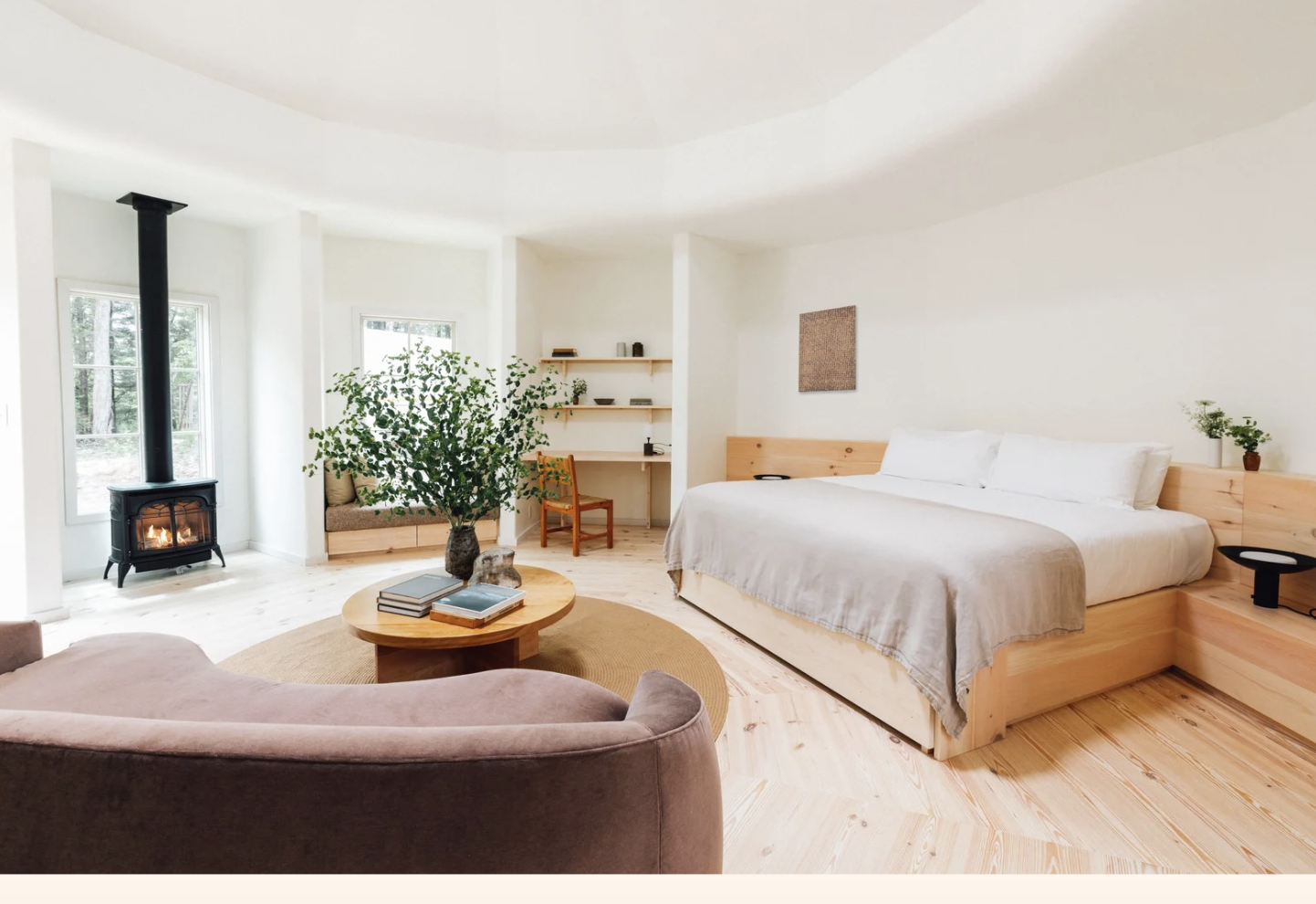 A cozy bedroom with a large bed, wooden platform, and white bedding. There is a small desk with a chair, a large display plant, a circular coffee table with books and rocks, and a windowed corner with a wood stove. The room has light wood floors and white walls.