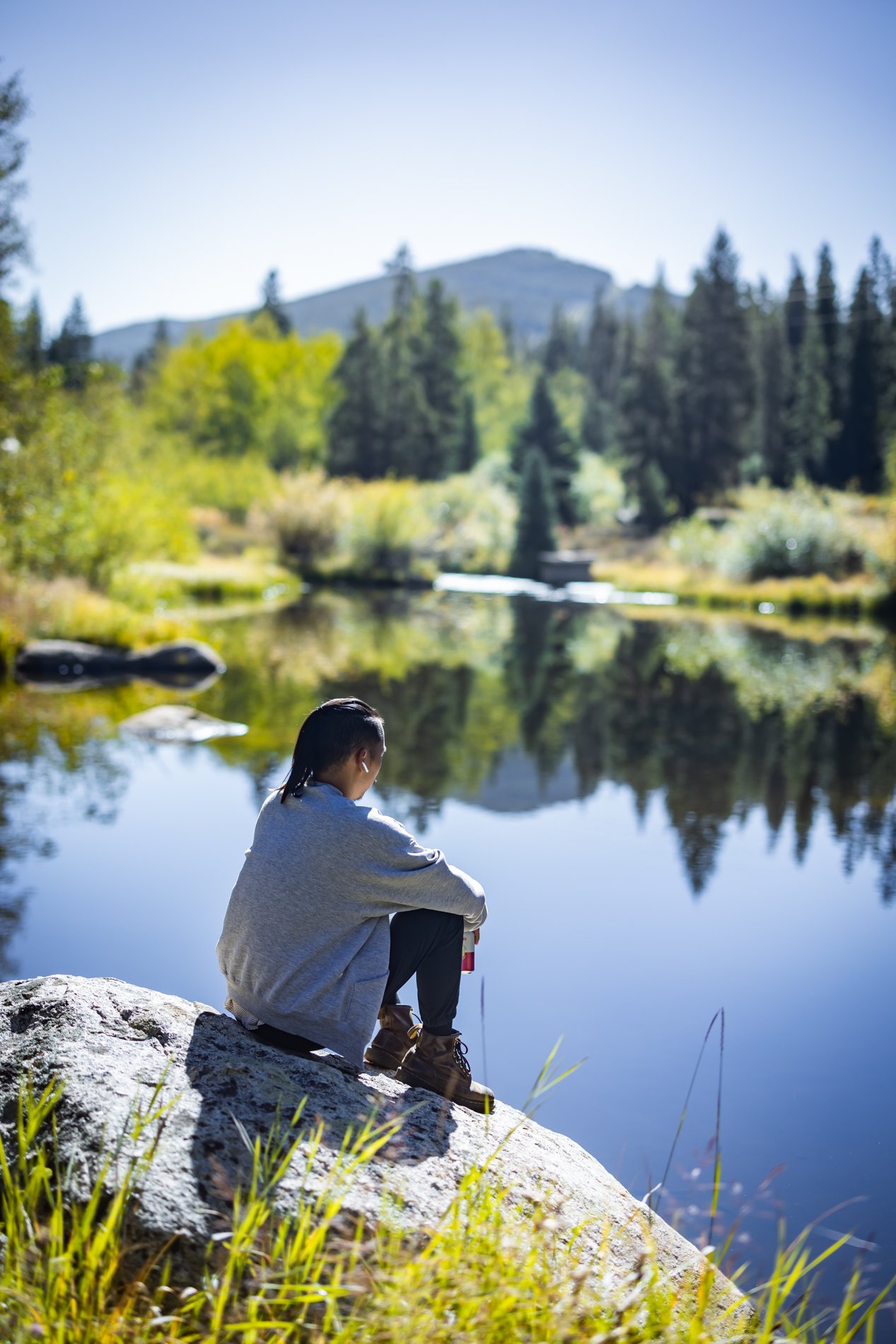 Person sitting on a rock by a calm river in a forested area with mountains in the background.