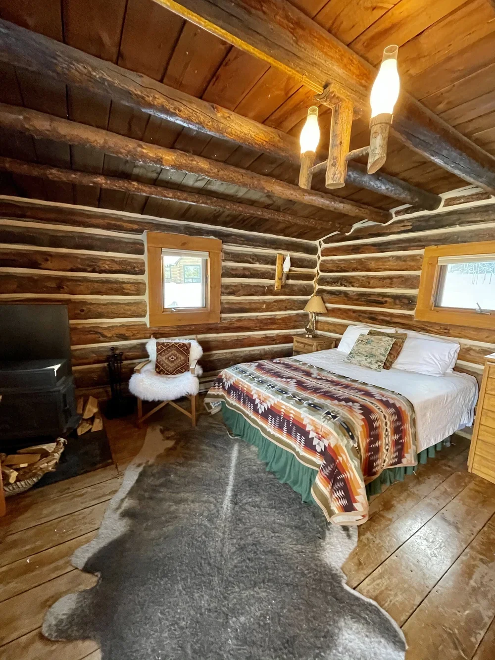A rustic log cabin bedroom with wood paneling on the walls and ceiling, featuring a bed with patterned bedding and pillows, small wooden nightstands, a chair with a furry cover, a TV, and a cowhide rug on the wooden floor.