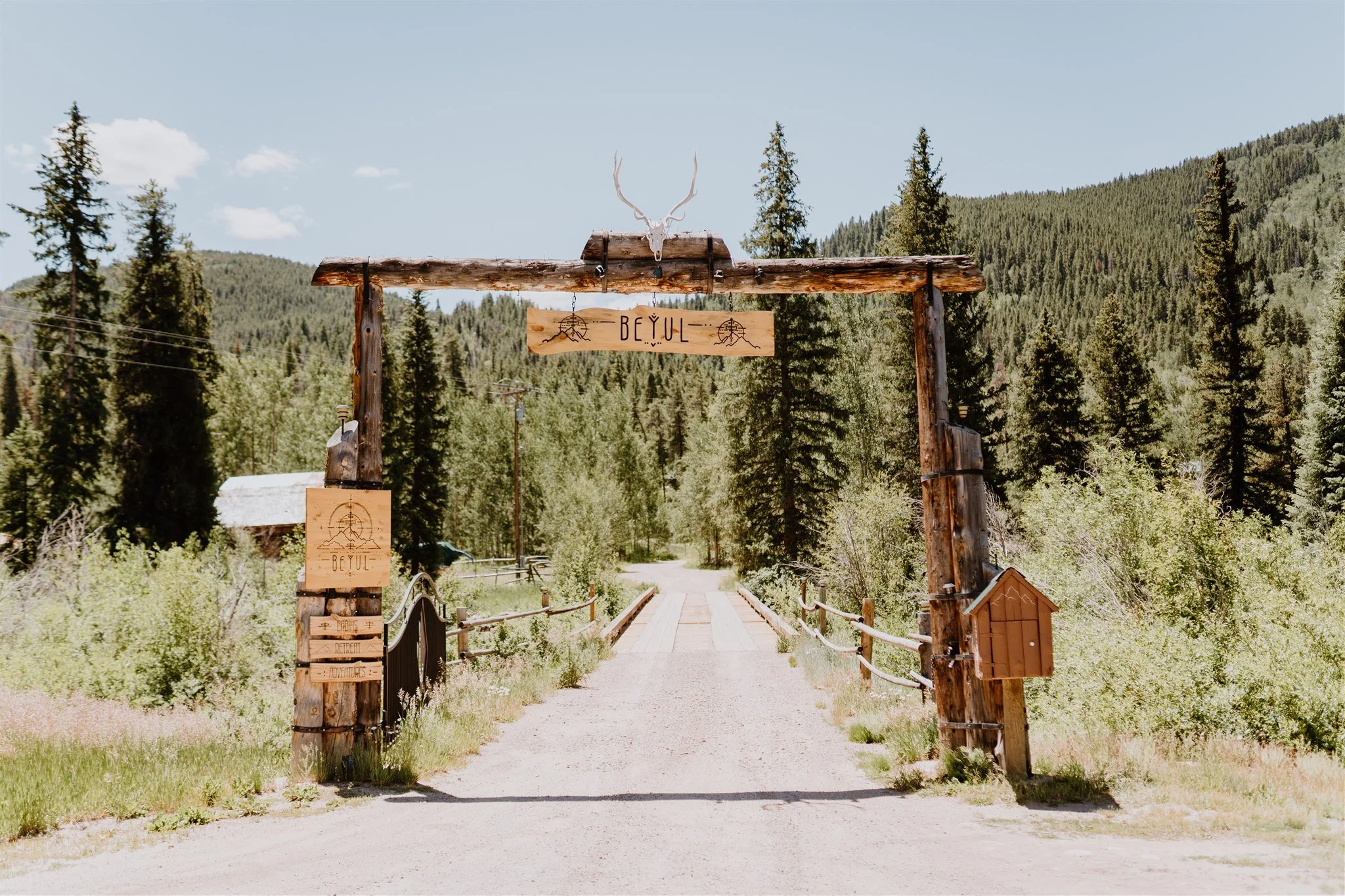 Rustic wooden gate with deer antlers and a sign that reads 'BEYUL,' leading into a forested area with mountains in the background.