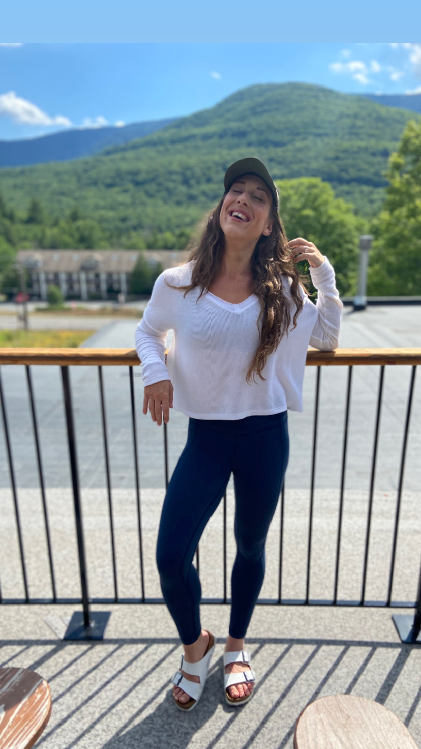 A woman in a white sweater, navy pants, and white sandals, standing on a balcony with mountains and green trees in the background, smiling with eyes closed and one hand on her hair.