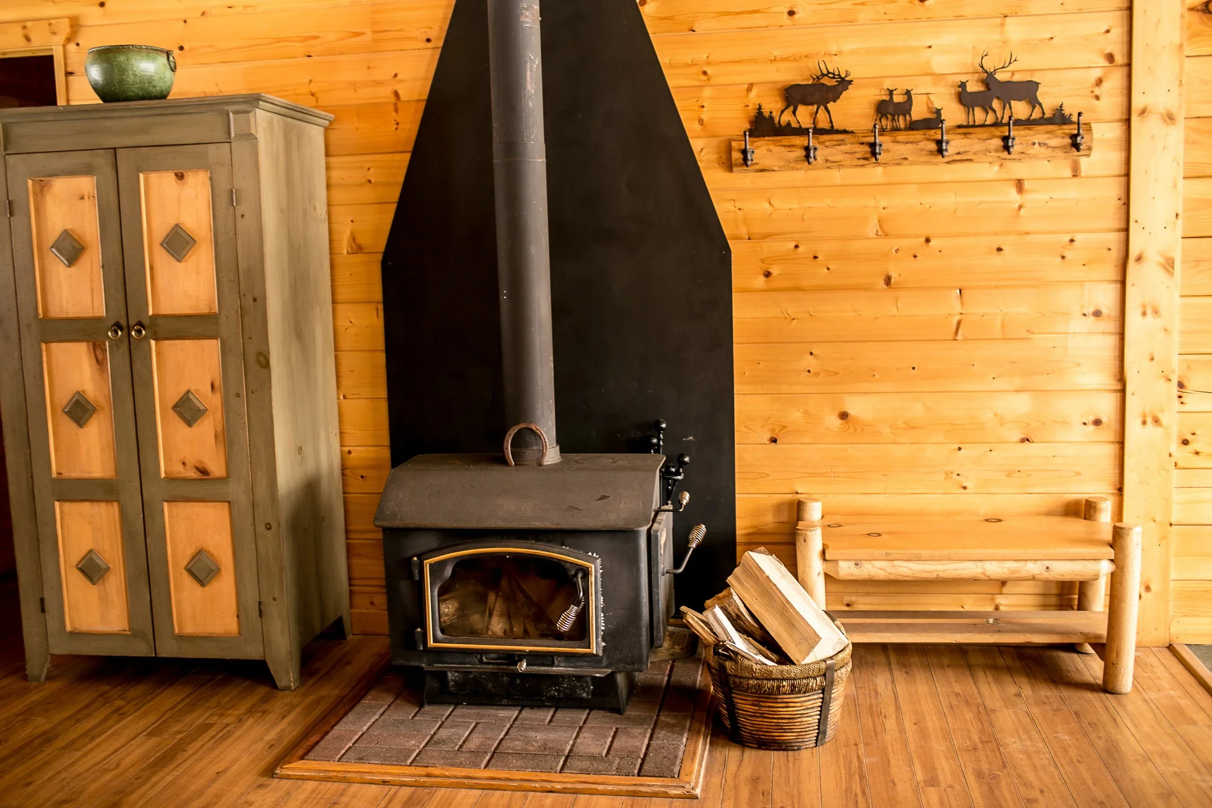 Interior of a wood cabin with a wood-burning stove, a basket of firewood, a wooden bench, a green vase on a cabinet, and a wall decoration of metal elk and deer silhouette hooks.