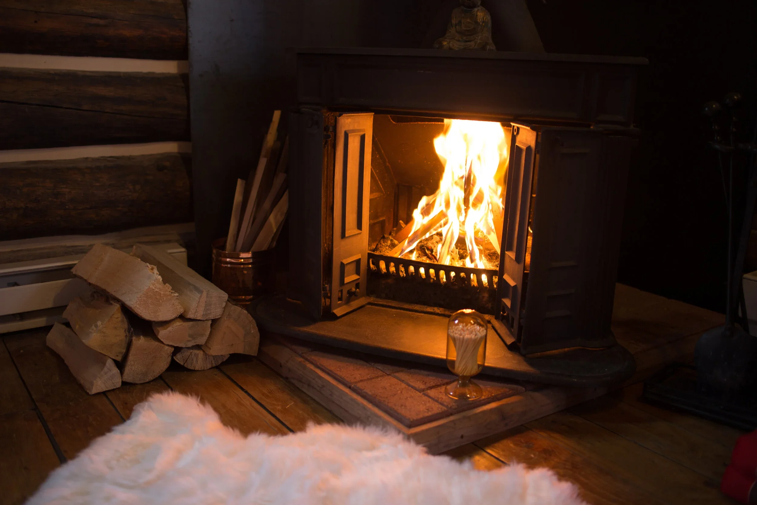 A warm fireplace with a roaring fire inside a cozy rustic cabin, surrounded by wood logs and a sheepskin rug.