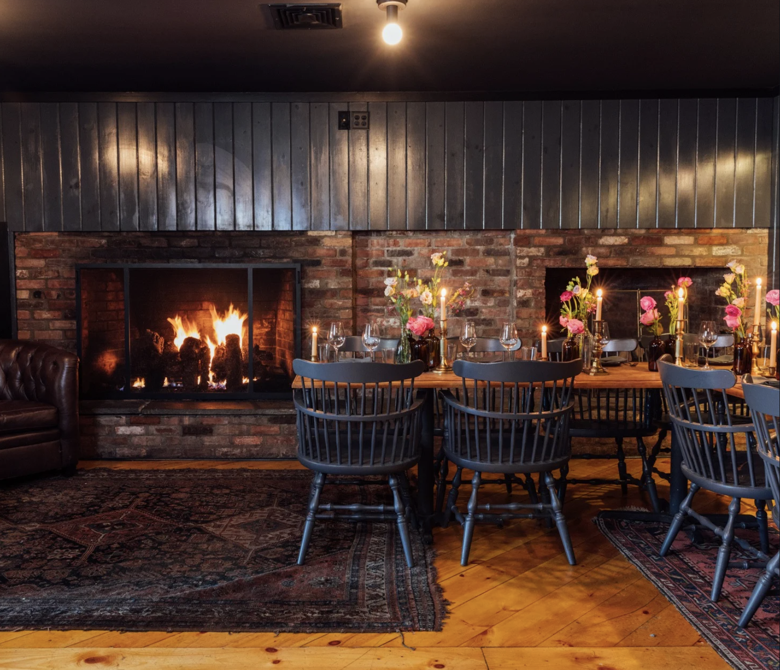A cozy dining area with a brick fireplace, a wooden dining table set with wine glasses, pink flowers, candles, and black chairs, located in a rustic room with dark wood wall paneling and a patterned rug.