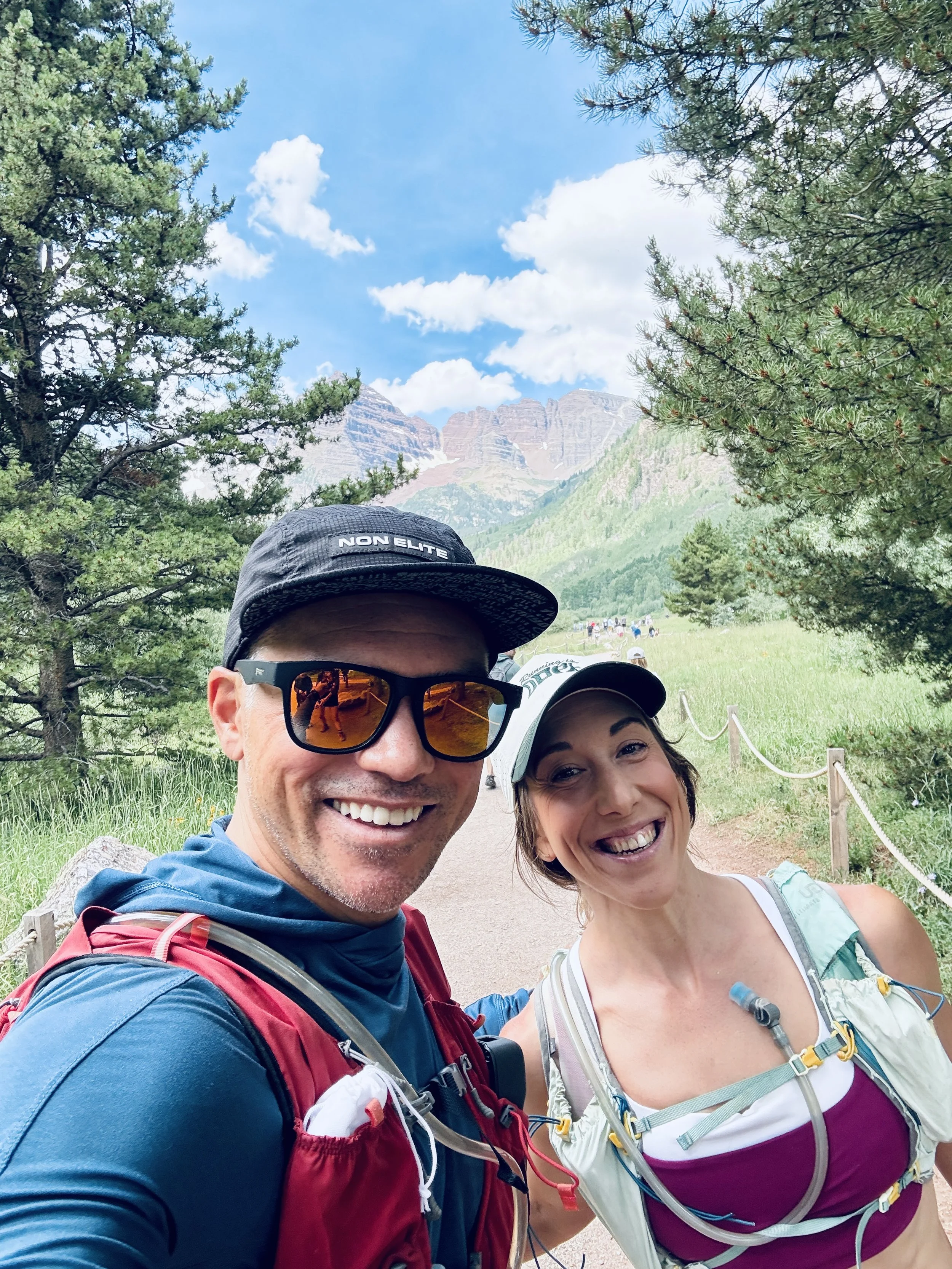 Smiling man and woman taking a selfie outdoors on a mountain trail surrounded by trees and green mountains with blue sky and clouds in the background.