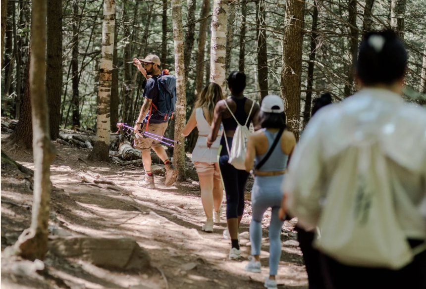 A group of people hiking through a forest on a trail, led by a man with trekking poles.