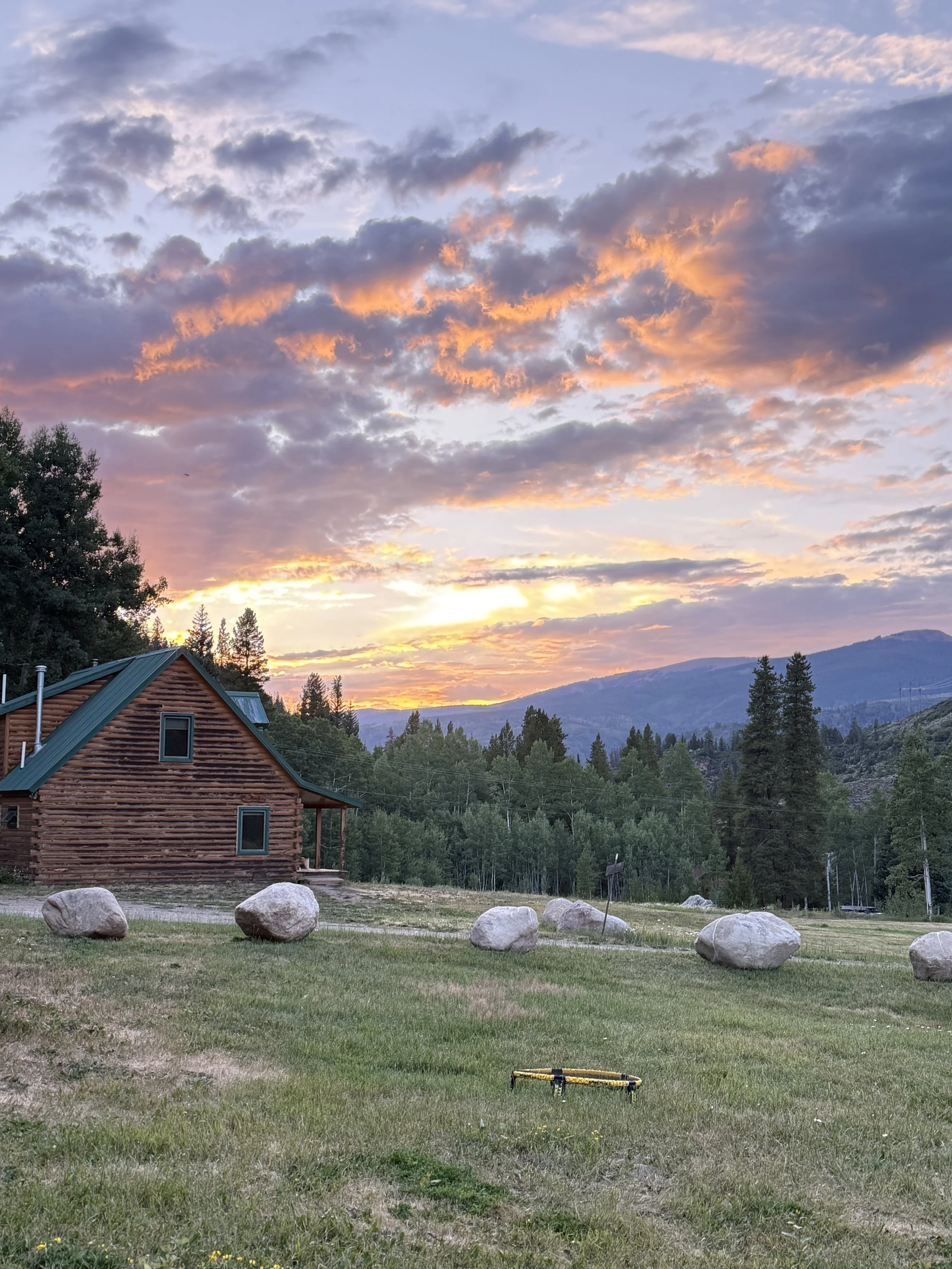 A cabin with a green metal roof in a grassy field, with large rocks in the foreground, surrounded by trees and mountains under a colorful sunset sky with clouds.
