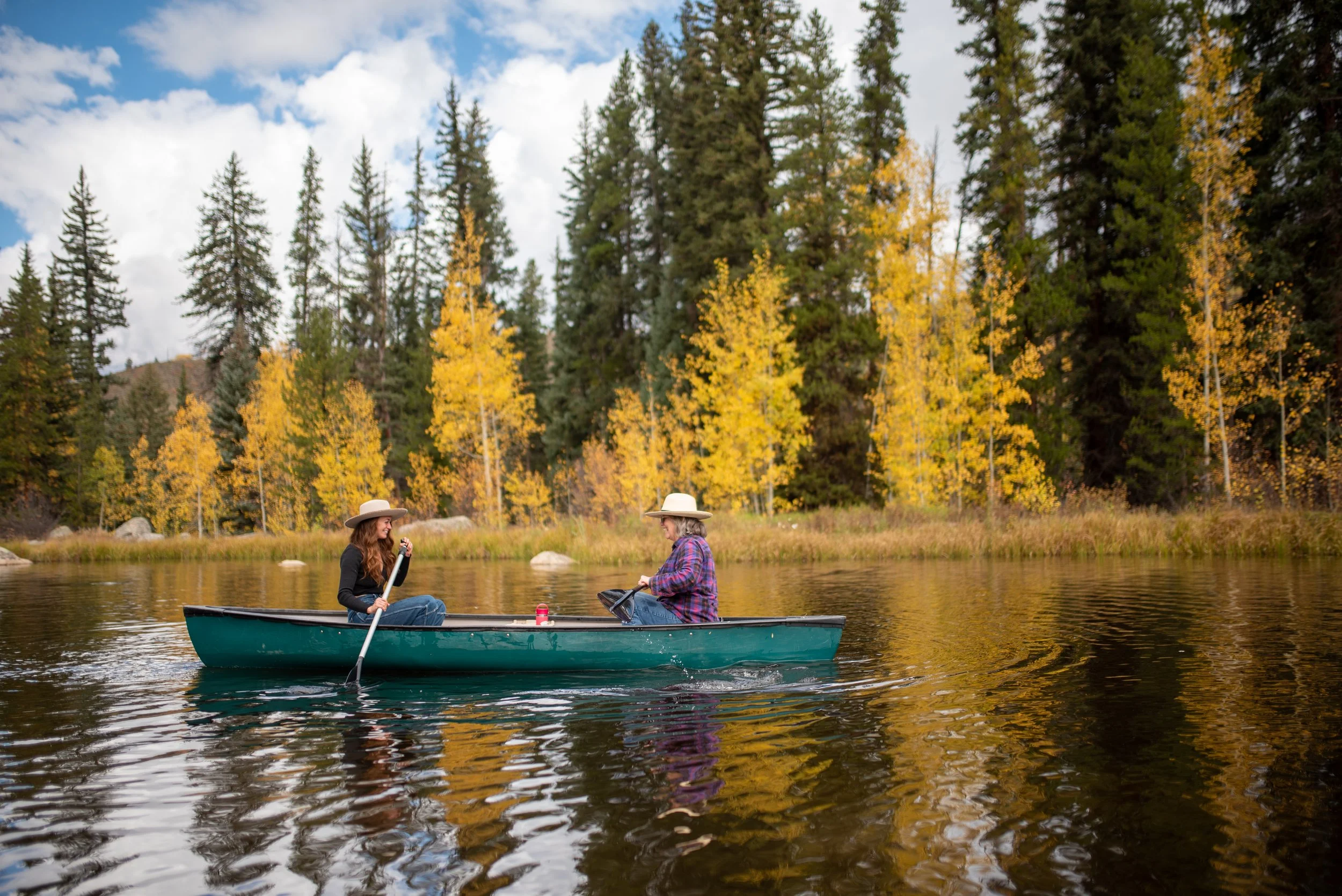 Two women wearing hats and plaid shirts enjoying a boat ride on a lake surrounded by autumn-colored trees.