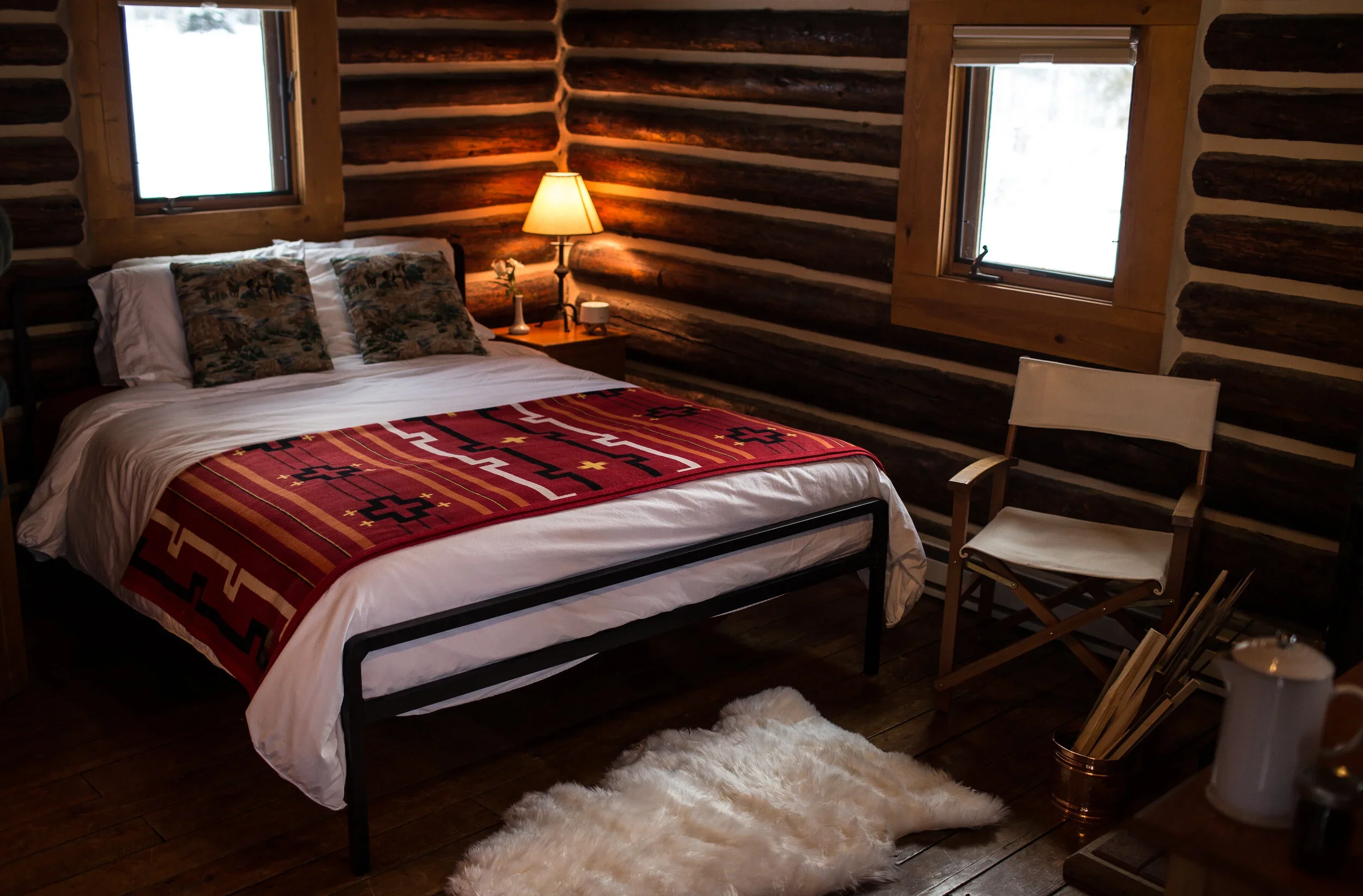 A cozy bedroom with rustic log walls, a neatly made bed with white linens, decorative pillows, and a southwestern-patterned blanket. A bedside table has a lamp and a small vase. Two windows let in natural light. A white director's chair and a fluffy white rug on a wooden floor complete the room's warm, inviting atmosphere.