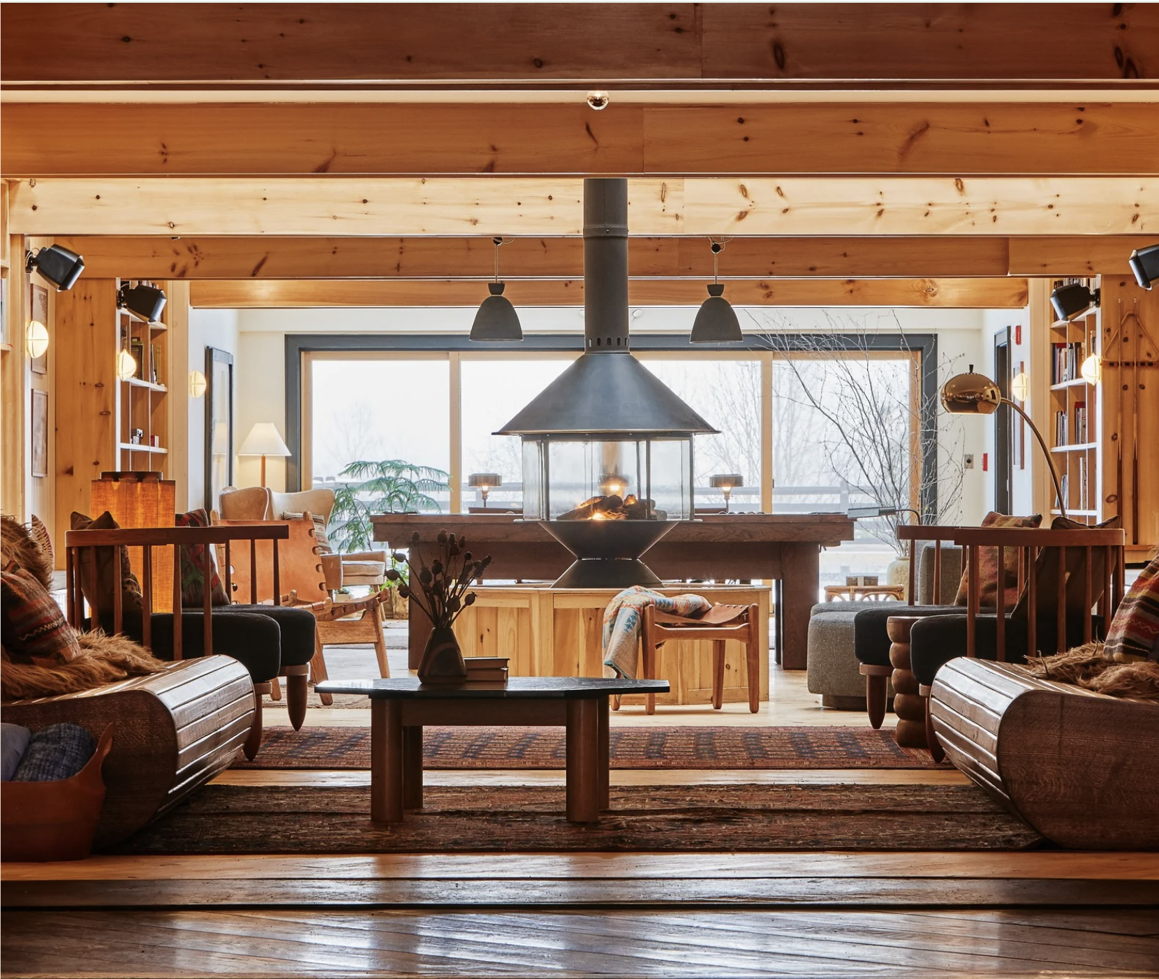 A cozy living room with wooden walls and ceiling, large windows, a fireplace, and comfortable seating.