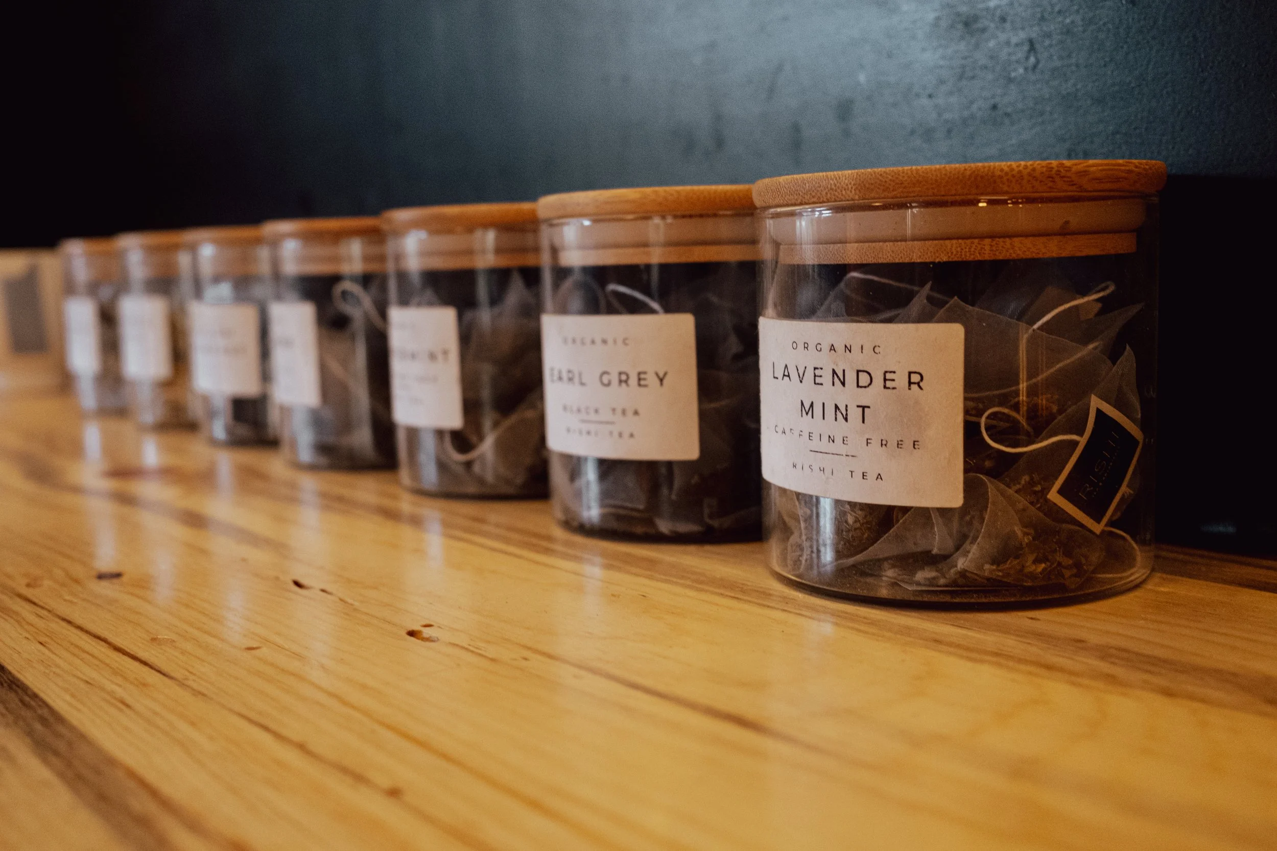 Clear containers with wooden lids containing various loose leaf teas lined up on a wooden surface, with labels indicating different flavors including lavender mint and Earl Grey