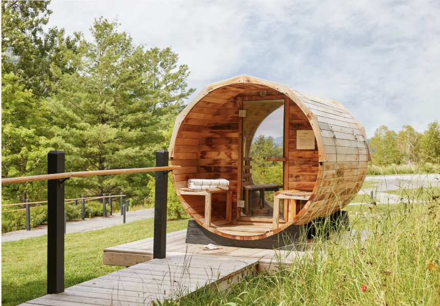 A wooden barrel-shaped sauna with an open door, situated in a lush green outdoor setting with trees and a pathway nearby.