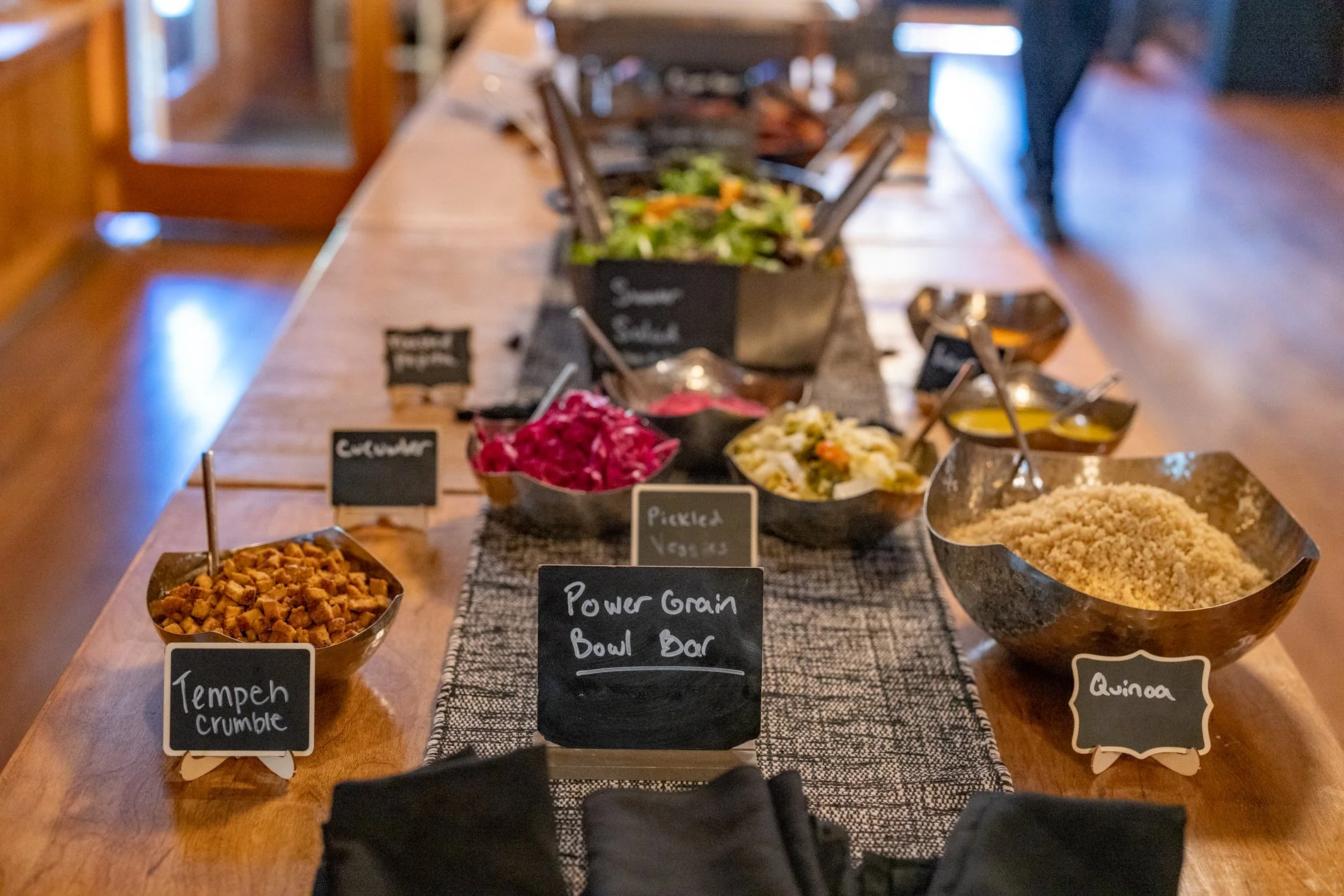 Salad bar setup with bowls containing various ingredients such as tempeh crumble, cabbage, quinoa, and pickled vegetables, labeled with small chalkboard signs, on a wooden table in a rustic setting.